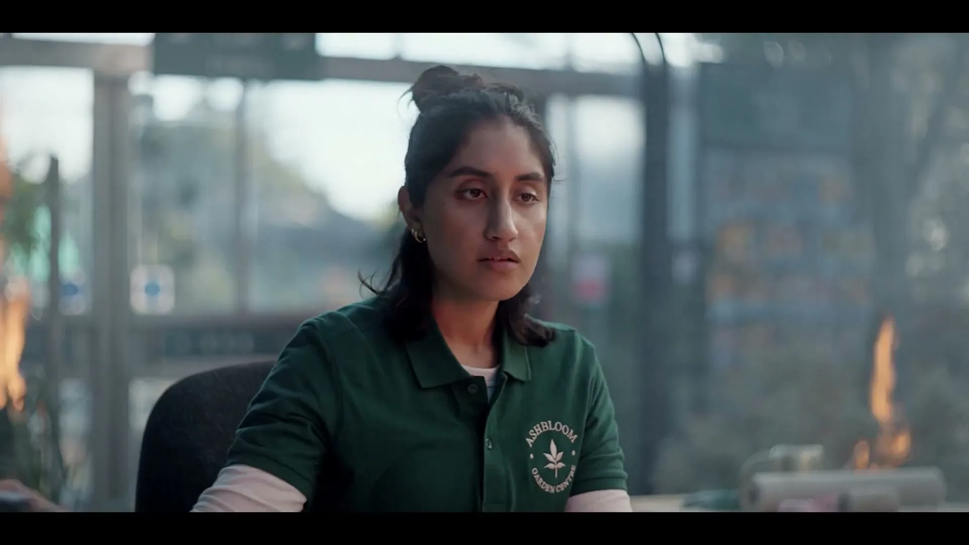 A young woman with dark hair tied up, wearing a green polo shirt, sits at a desk in a garden center, looking around with concern as fires burn in the background.