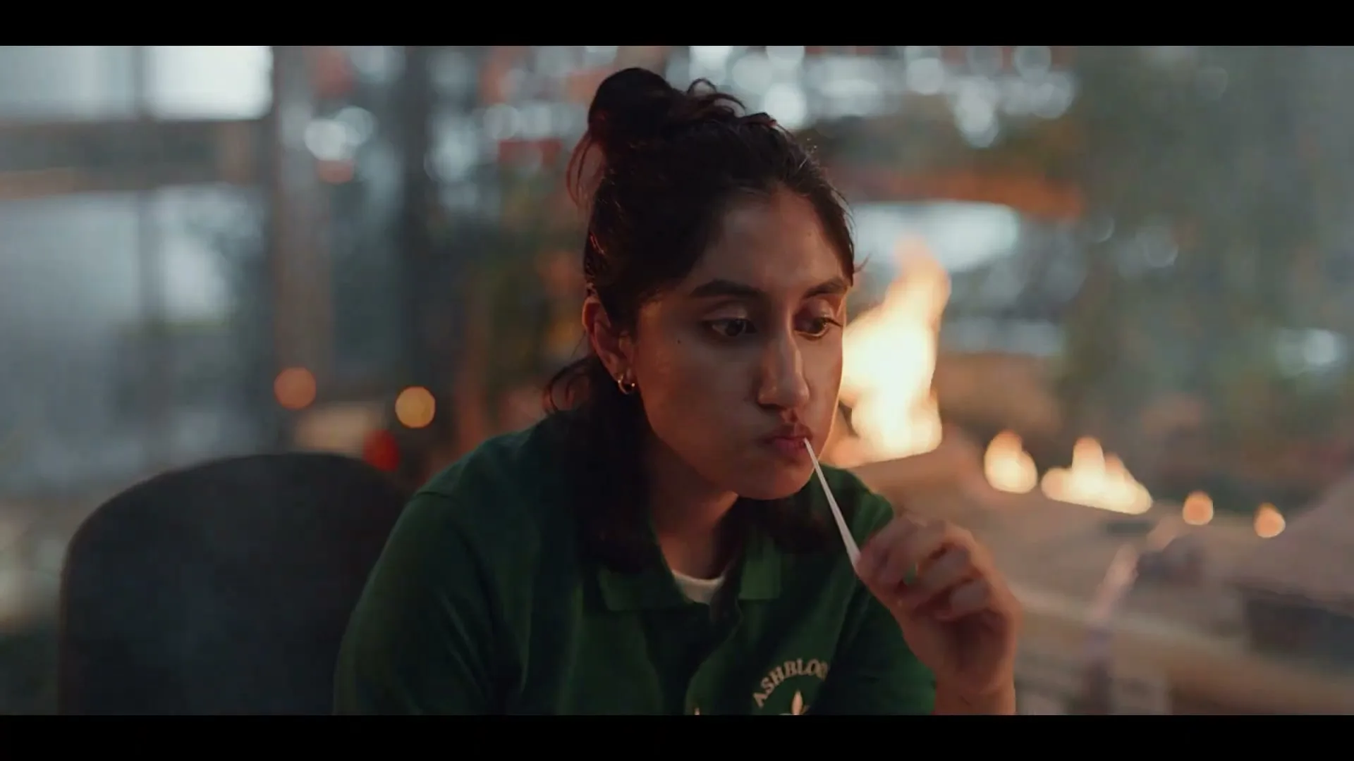A young woman with dark hair in a bun, wearing a green polo shirt, sits by a campfire and pulls a melted marshmallow from a stick.