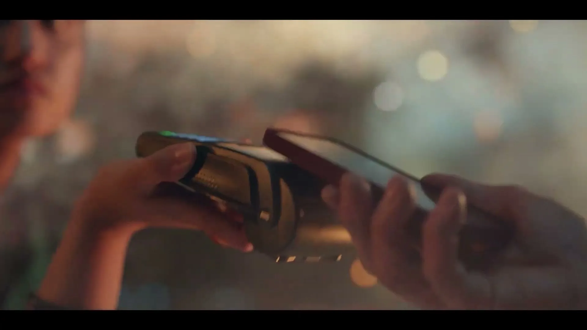A person holds a payment terminal while another person taps their phone to it, completing a transaction in a dimly lit, blurry background.