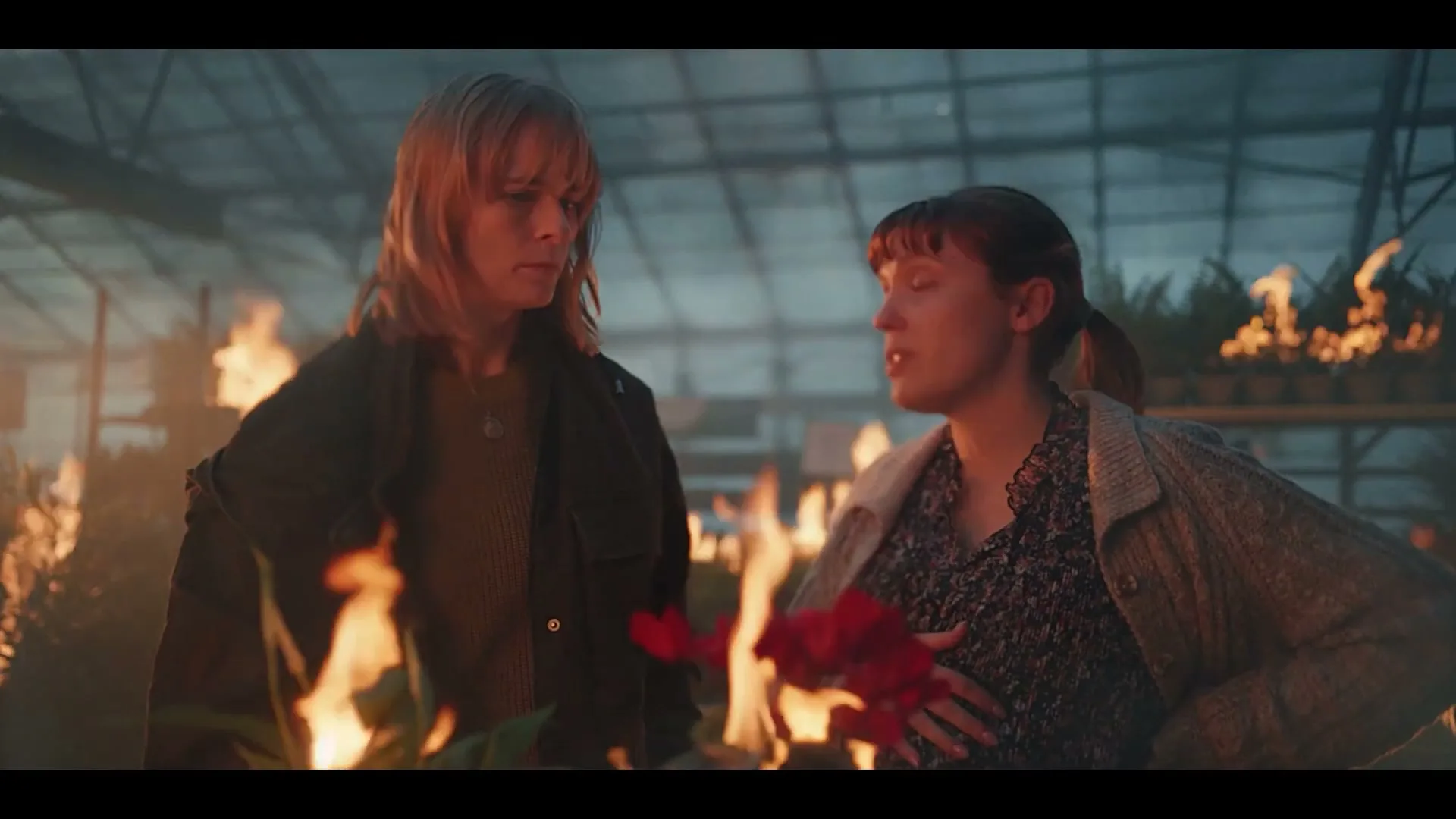 Two women stand in a burning greenhouse, surrounded by flaming plants, as one woman expresses indecision.