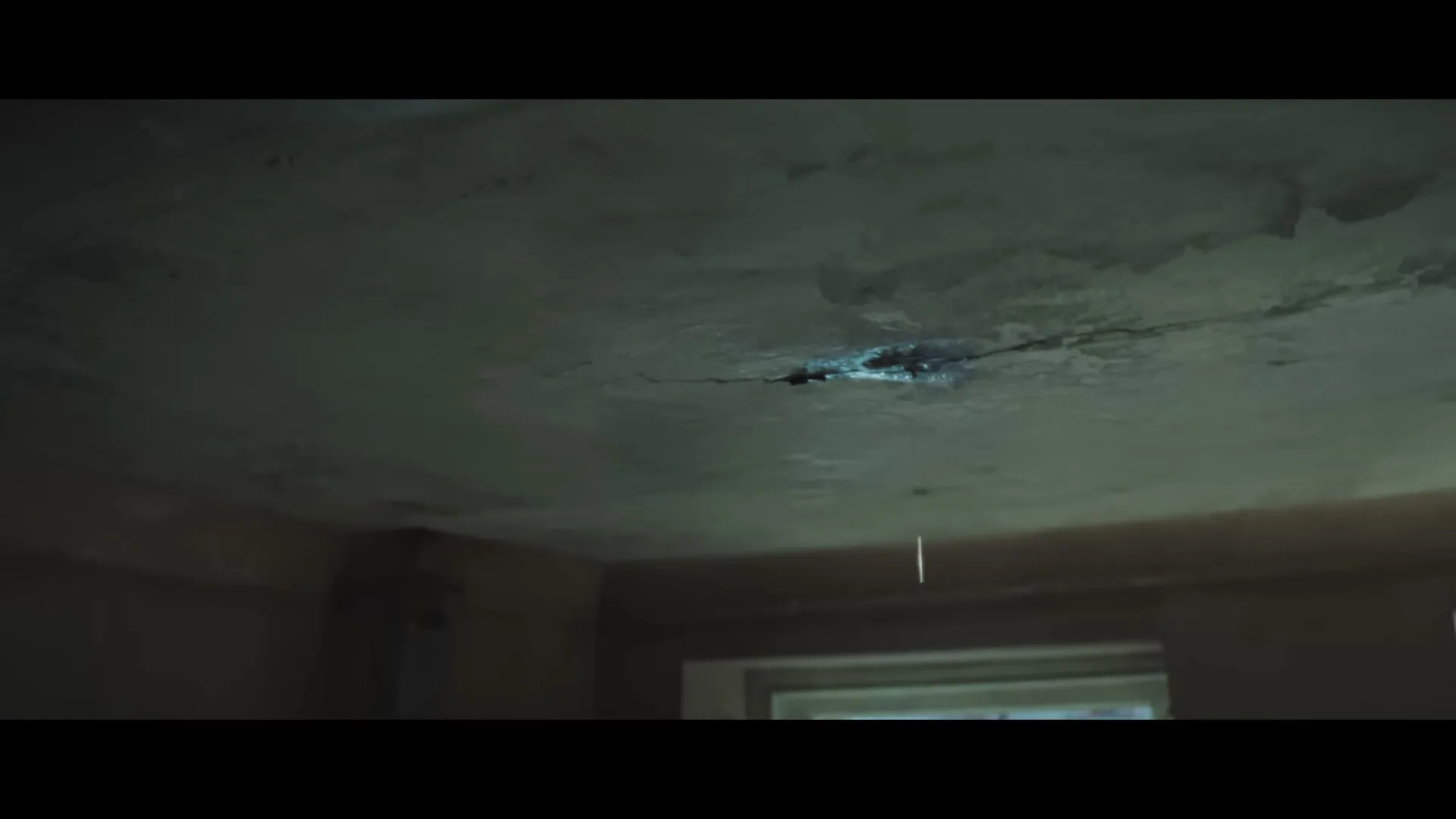 A dark, cracked ceiling with a glowing blue hole in the center, from which water drips, in an empty room.