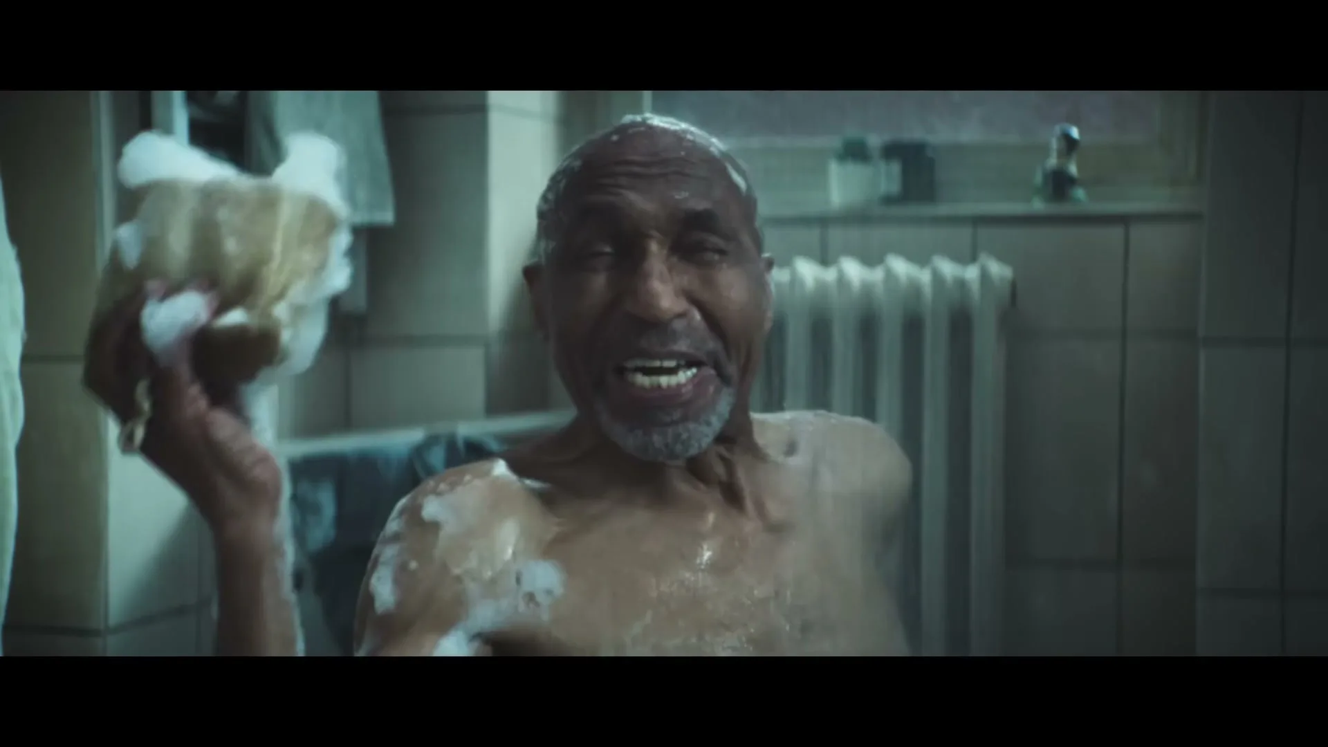 A man with a white beard is in a shower, covered in soap, holding a sponge, and smiling at the camera.