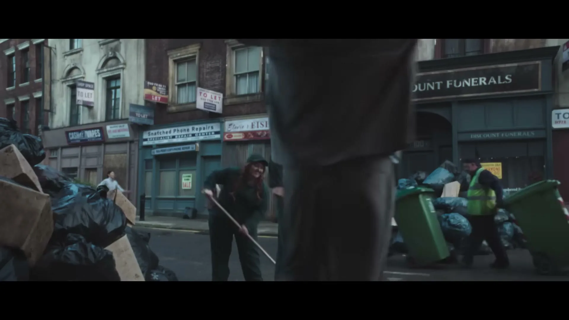 A man in a suit walks past a street filled with overflowing trash bags and bins, as sanitation workers sweep the street in front of "Derek's Discount Funerals."