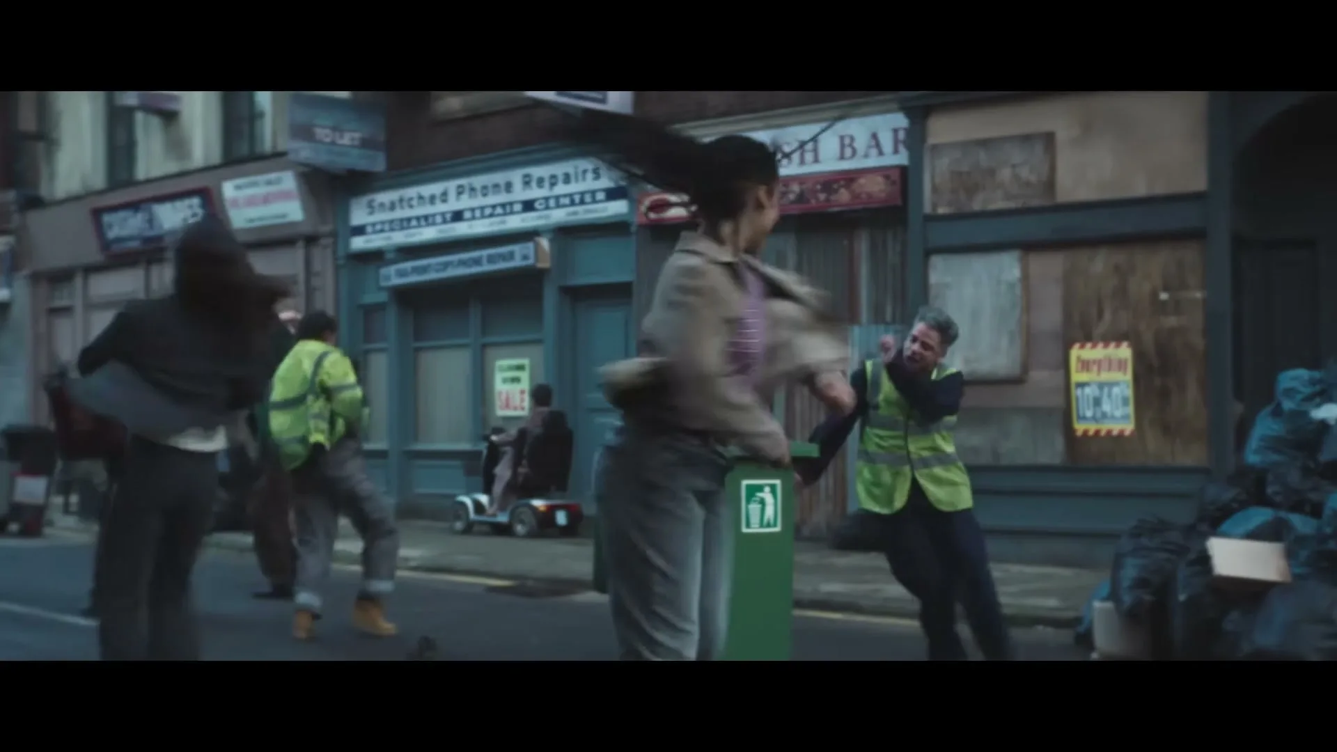 A diverse group of people in casual clothes dance erratically in a street filled with trash bags and boarded-up shops, with a green trash can in the foreground.