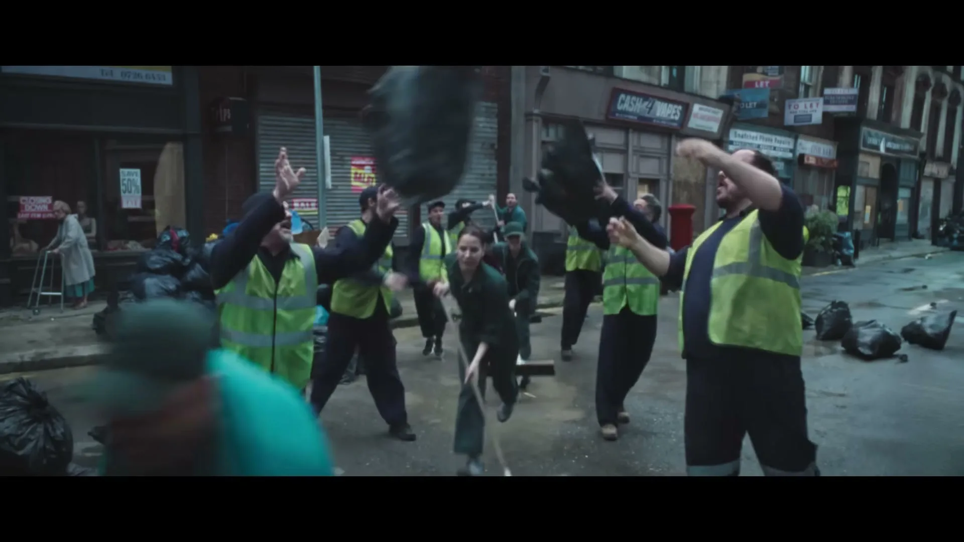 A group of sanitation workers in neon vests and dark uniforms are dancing and throwing trash bags in a dirty city street filled with garbage.