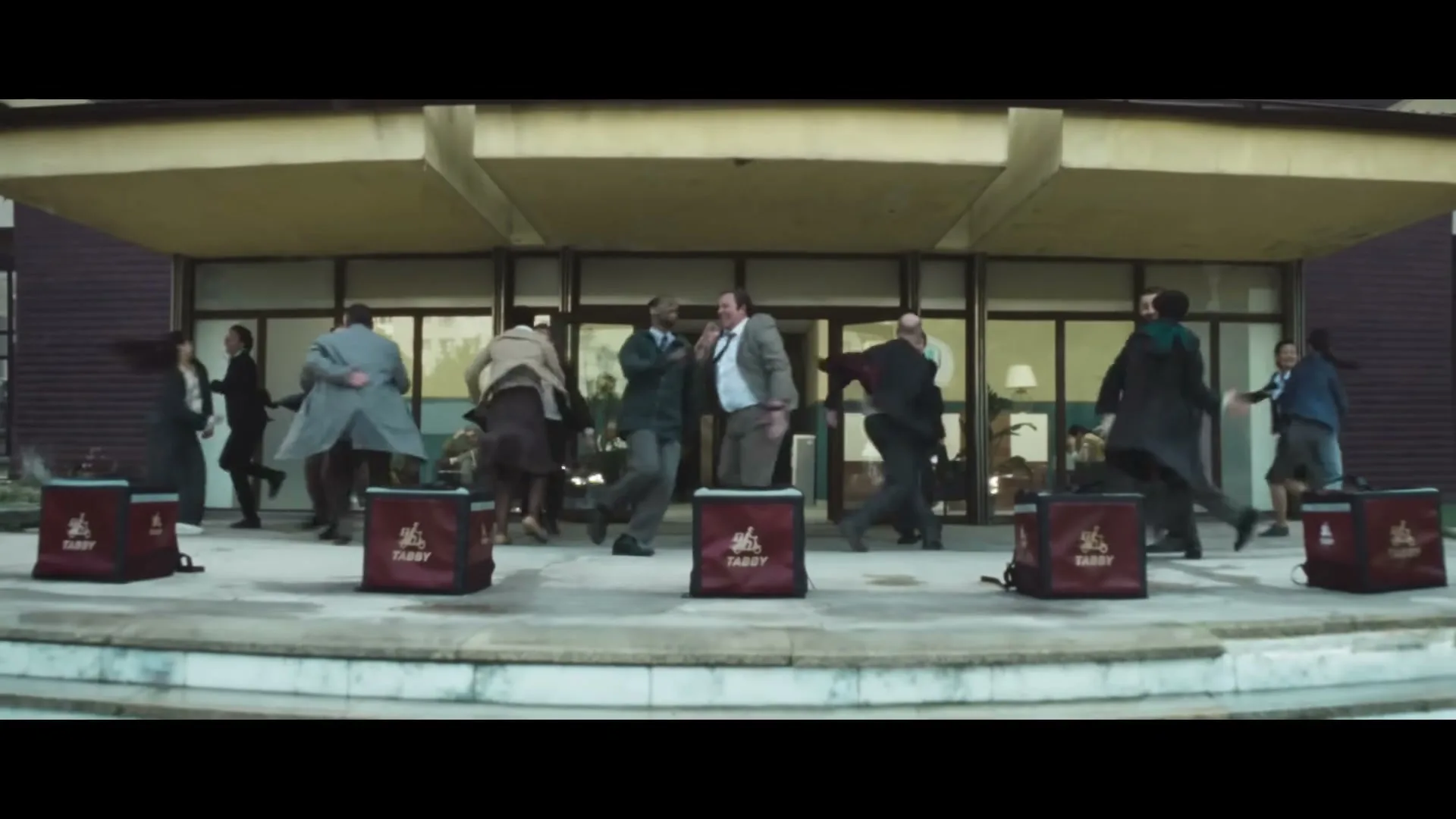 A diverse group of office workers in business attire dance energetically on a set of steps outside a modern building, with red delivery bags placed in front of them.