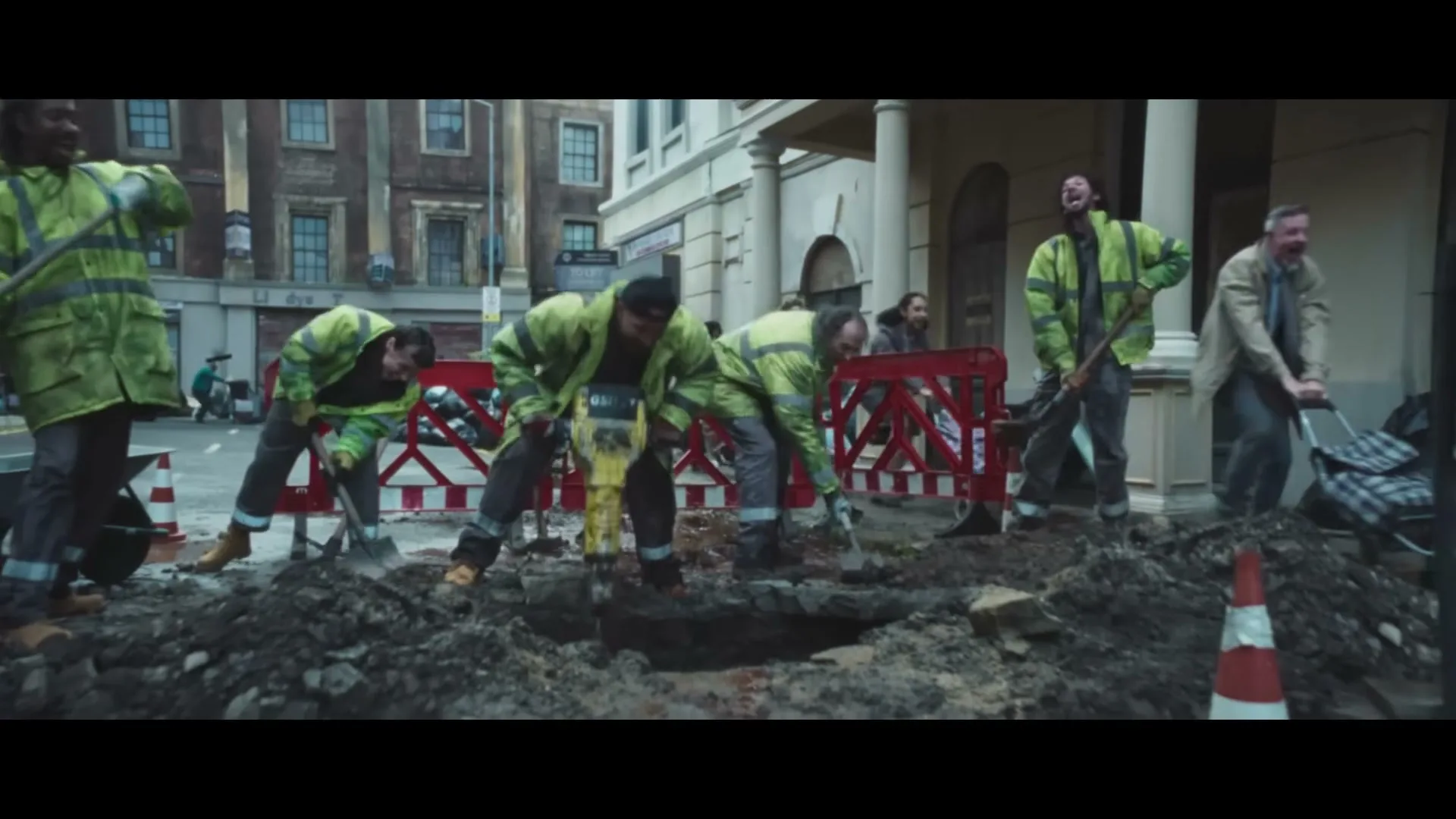 A group of construction workers in high-vis jackets are digging a hole in the street, as mud erupts from the ground.