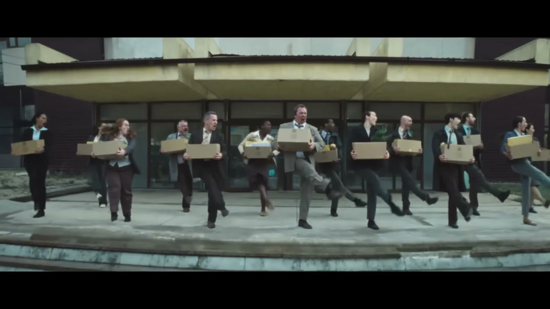 A diverse group of office workers in business attire, holding cardboard boxes, dance in unison outside a dilapidated building.