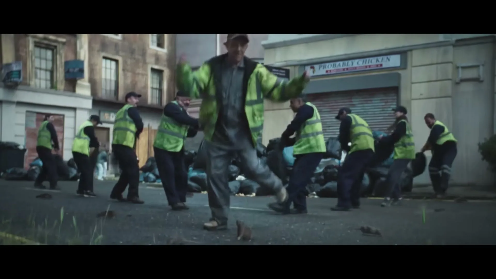 A group of sanitation workers in neon vests dance in the street, surrounded by piles of trash bags and rats, in an urban setting.