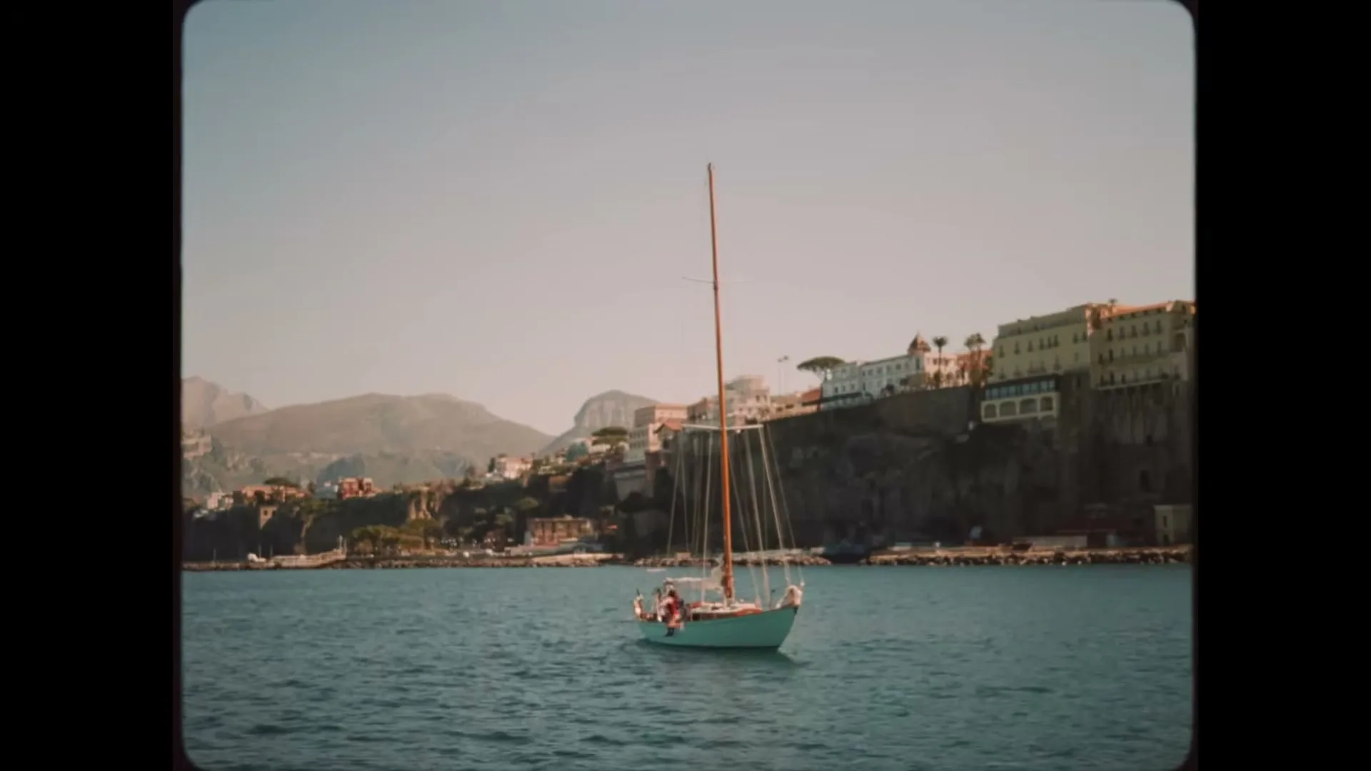 A sailboat with people on board sails across the blue sea, with a cliffside town and mountains in the background under a clear sky.