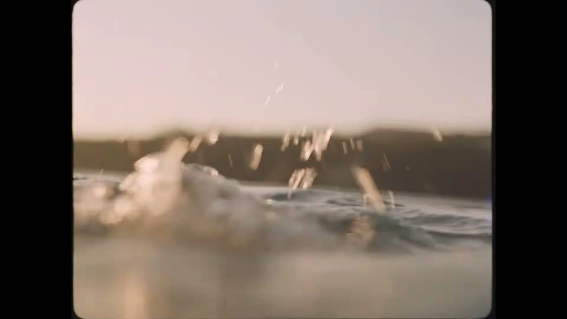 A low angle shot captures the water's surface as it splashes and ripples, with a blurry background of a distant shore under a bright sky.