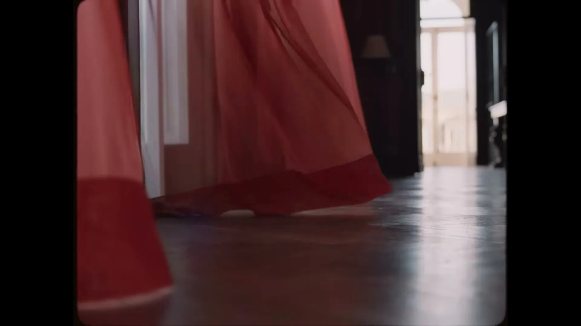 A person in a flowing red dress walks across a wooden floor in a dimly lit room with a bright doorway in the background.
