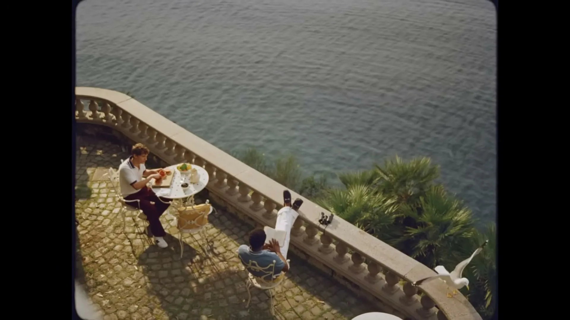 Two men relax on a sunny balcony overlooking the sea, one eating at a table, the other reading, as a seagull flies by.