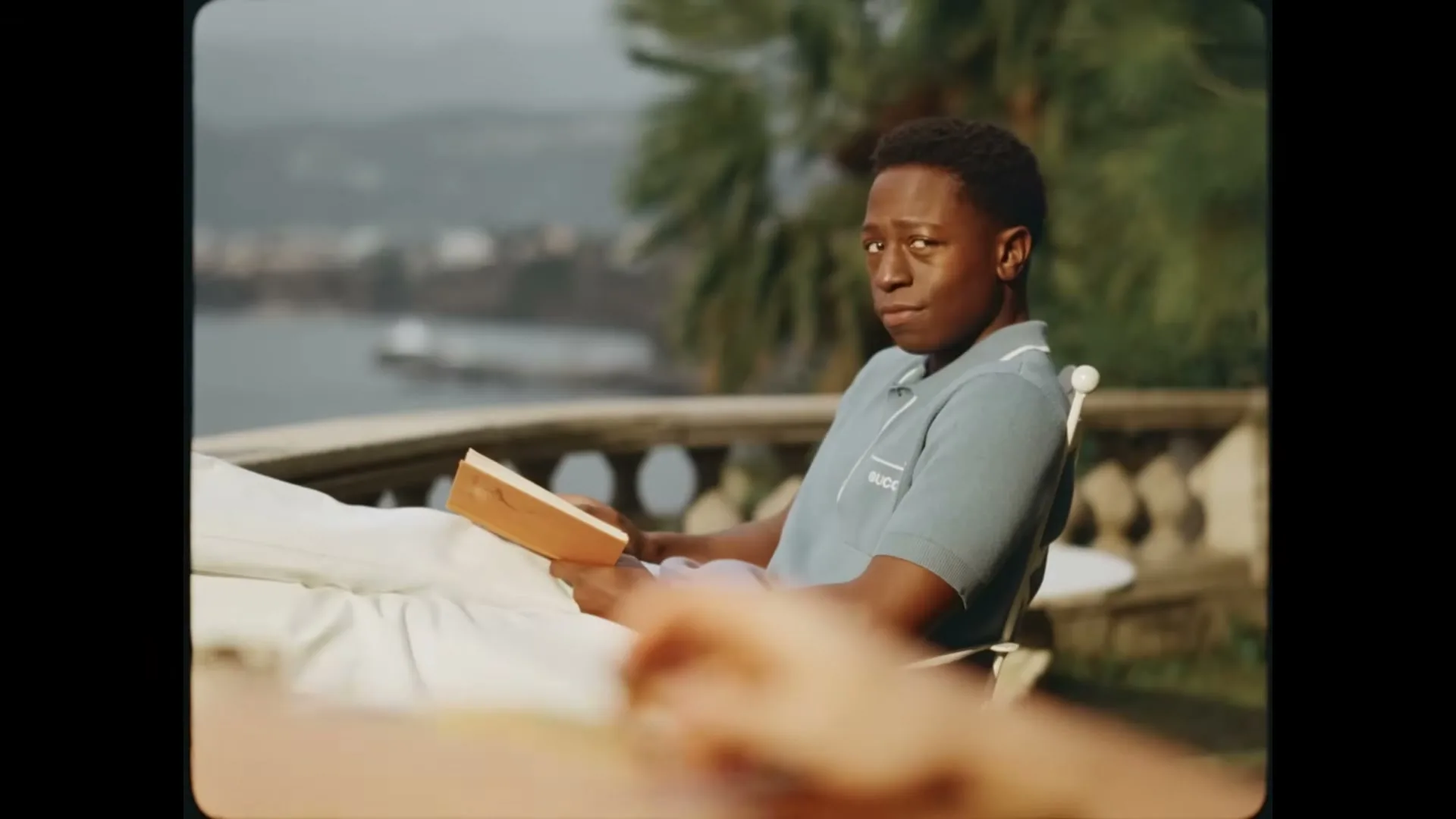 A young Black man in a light blue polo shirt reads a book on a sunny balcony overlooking the sea.