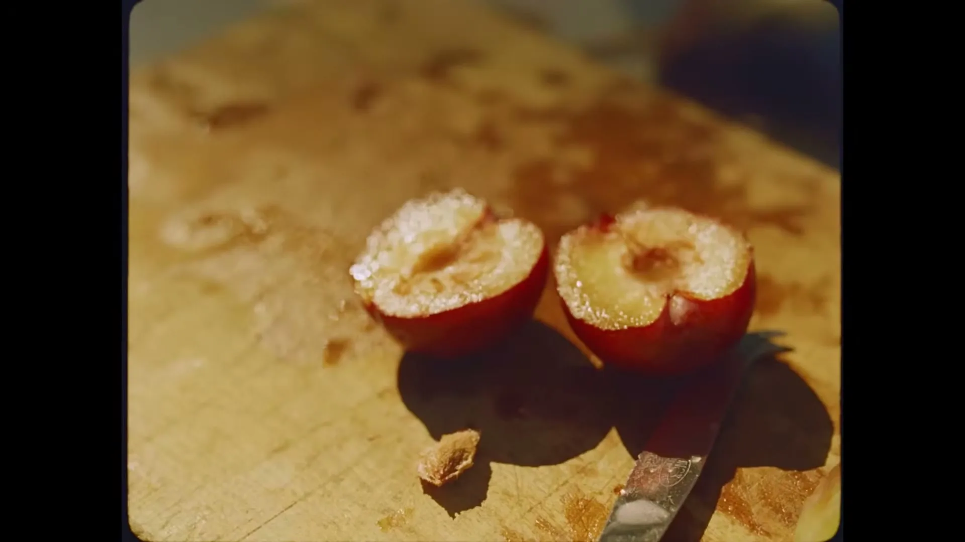 Two halves of a red plum, sprinkled with sugar, sit on a wooden cutting board next to a knife in a sunlit kitchen.