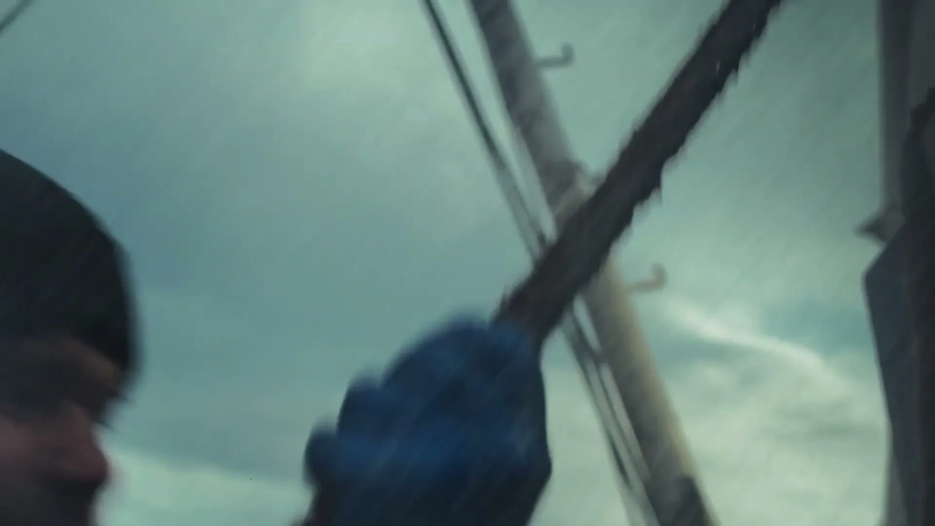 A male fisherman in a beanie and yellow jacket struggles with a rope on a boat in rough weather, with a cloudy sky in the background.
