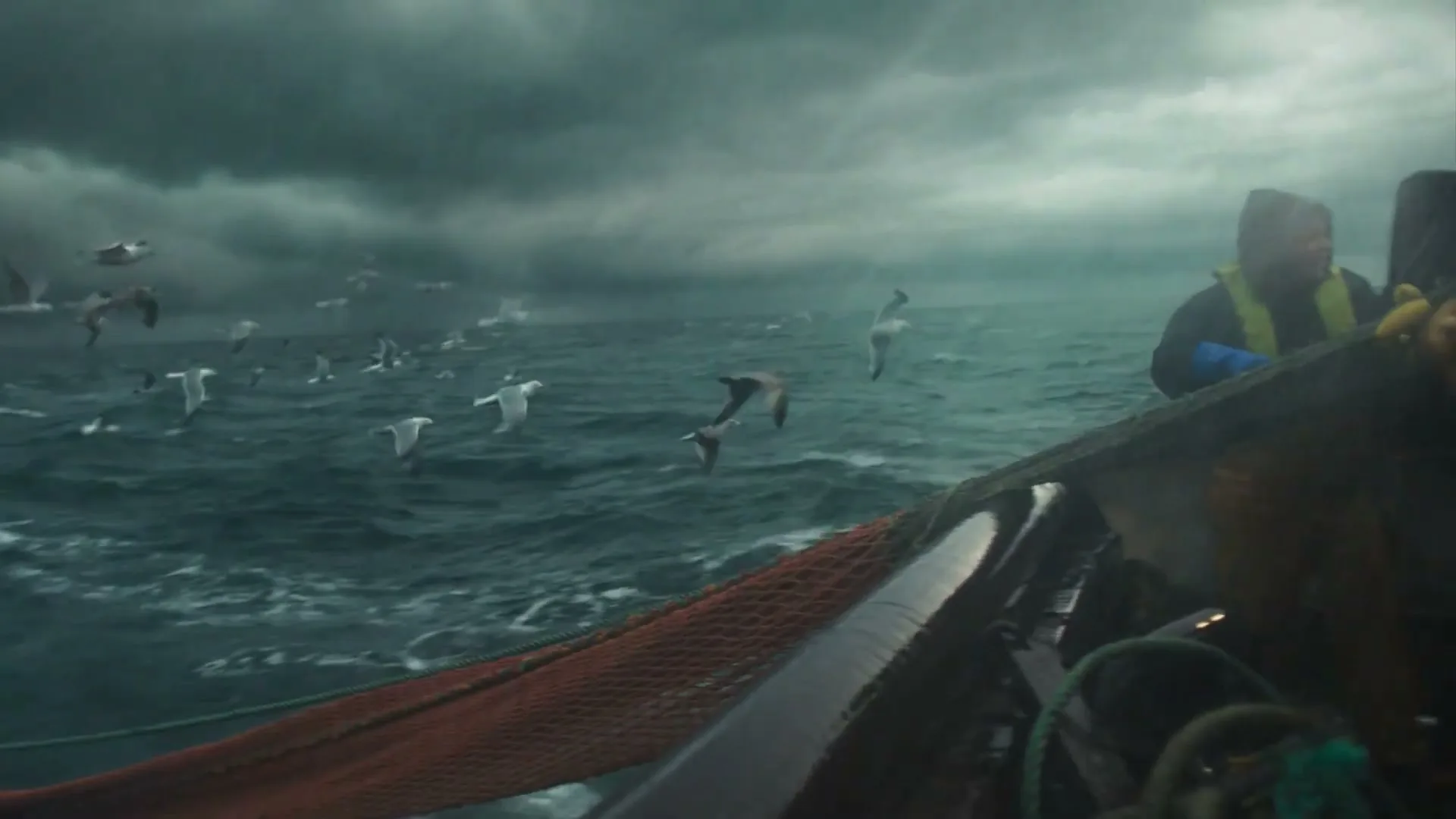 A fisherman in a yellow life vest and dark jacket stands on a boat, pulling in a red fishing net on a stormy, grey sea with many seagulls flying around.