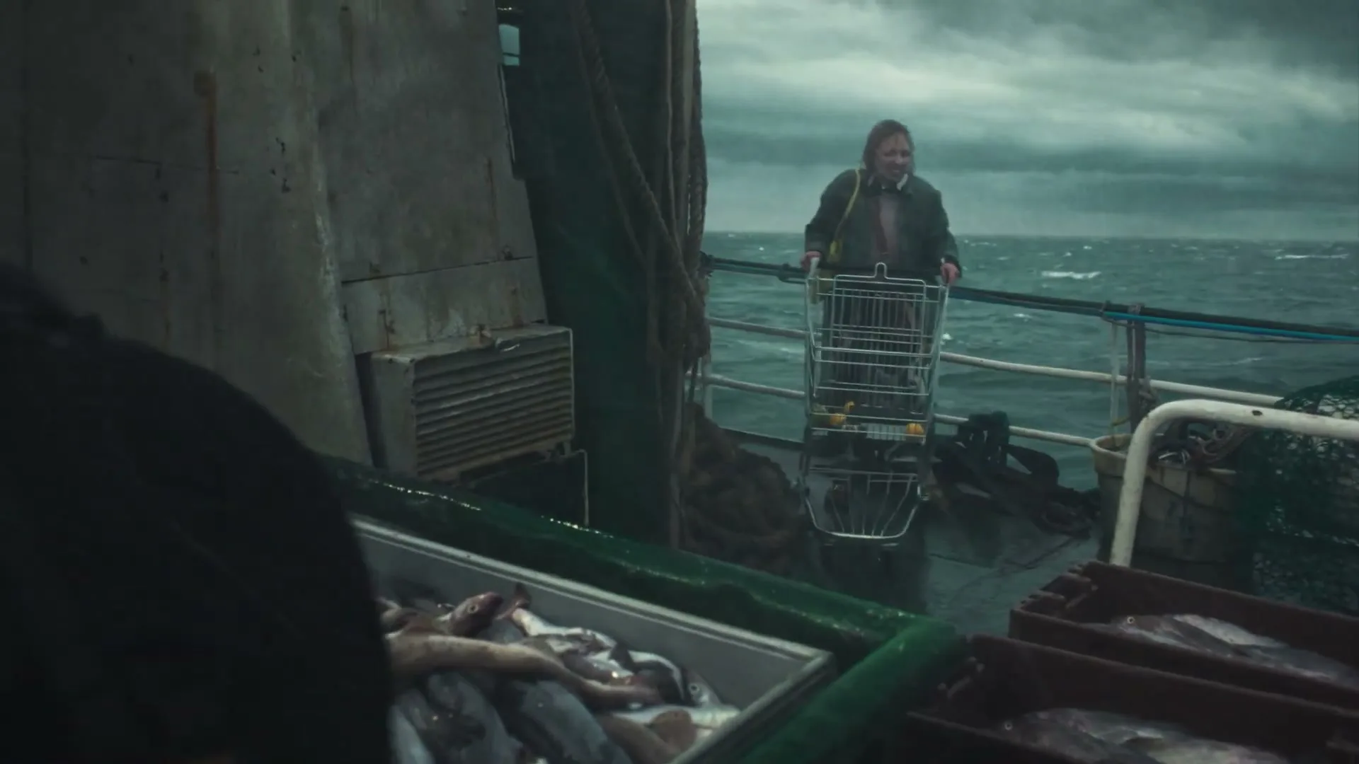 A female fisher in a green jacket pushes a shopping cart on a fishing boat, with crates of fish in the foreground and a stormy sea in the background.