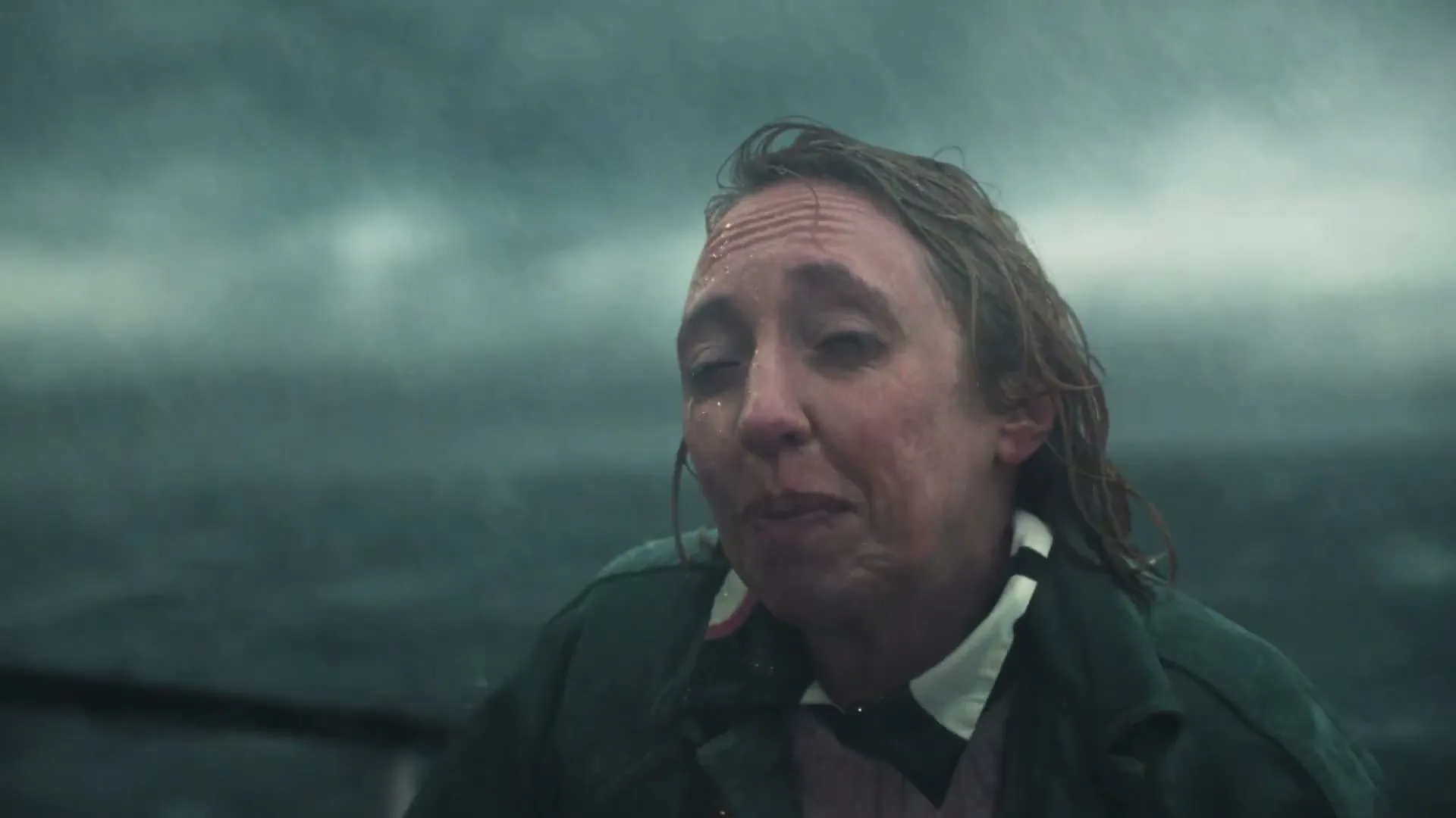 A woman in a green jacket and pink sweater, with wet hair and face, is on a boat in a stormy sea, making a peace sign and smiling.