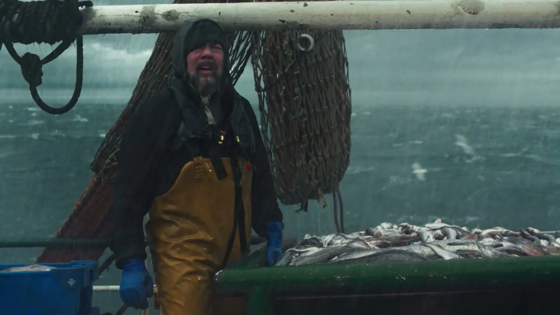 A male fisherman in yellow waders and a black jacket stands on a boat deck, looking tired, with a pile of fish beside him as rain falls and the sea churns in the background.