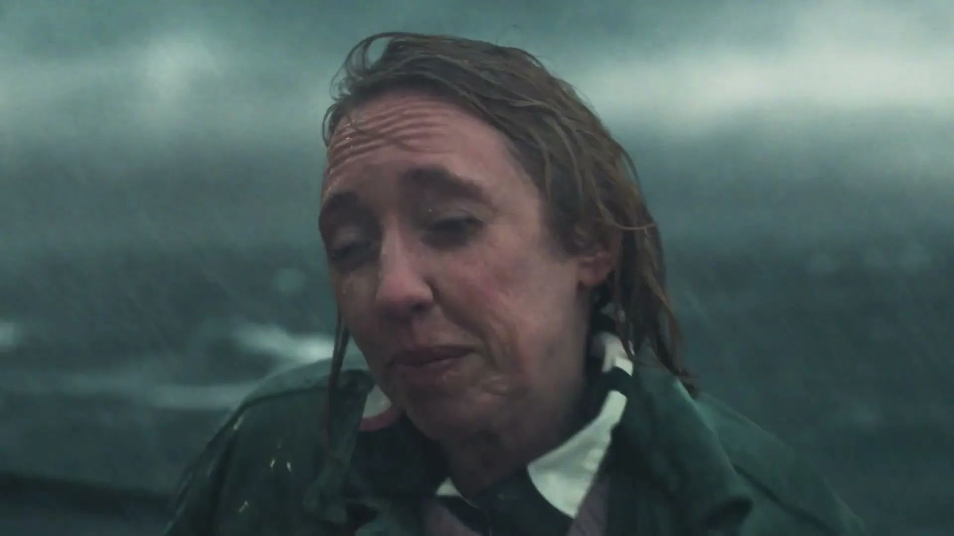A woman in a green jacket and white collar, with wet hair and face, looks distressed and speaks to the camera in a stormy, rainy outdoor setting.