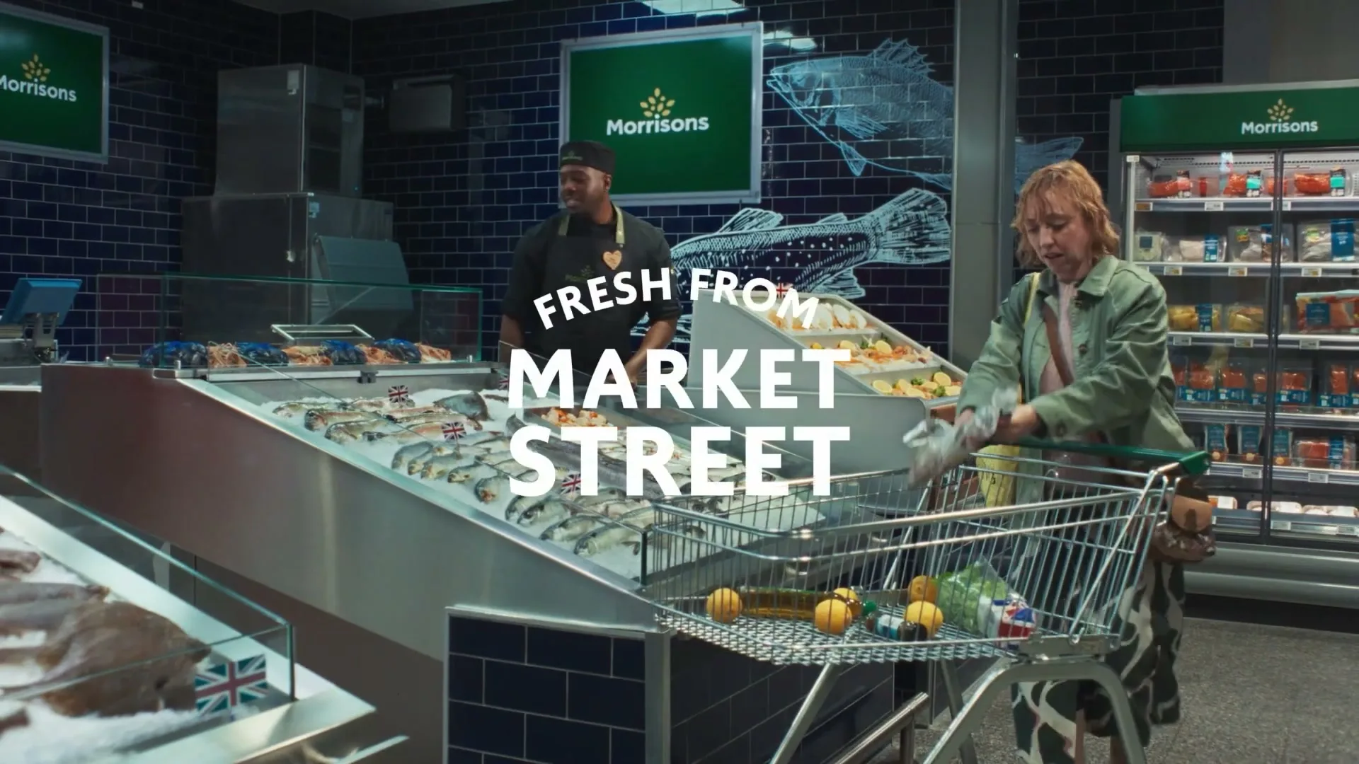 A male fishmonger hands a packaged fish to a female customer with a shopping cart in a brightly lit supermarket fish counter.