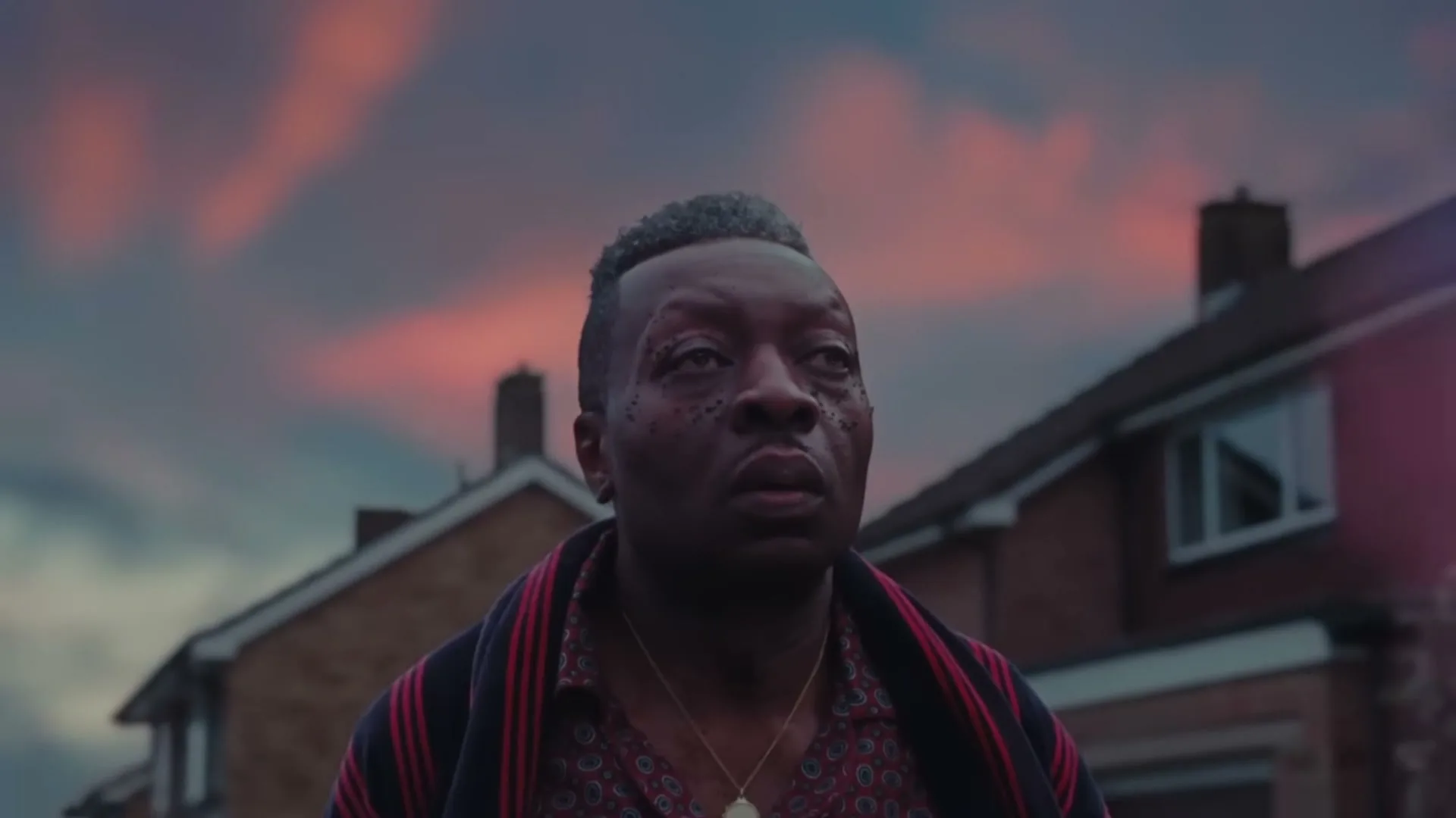 A Black man with a patterned shirt and striped robe looks up in wonder at something off-screen, with suburban houses and a dramatic sky in the background.