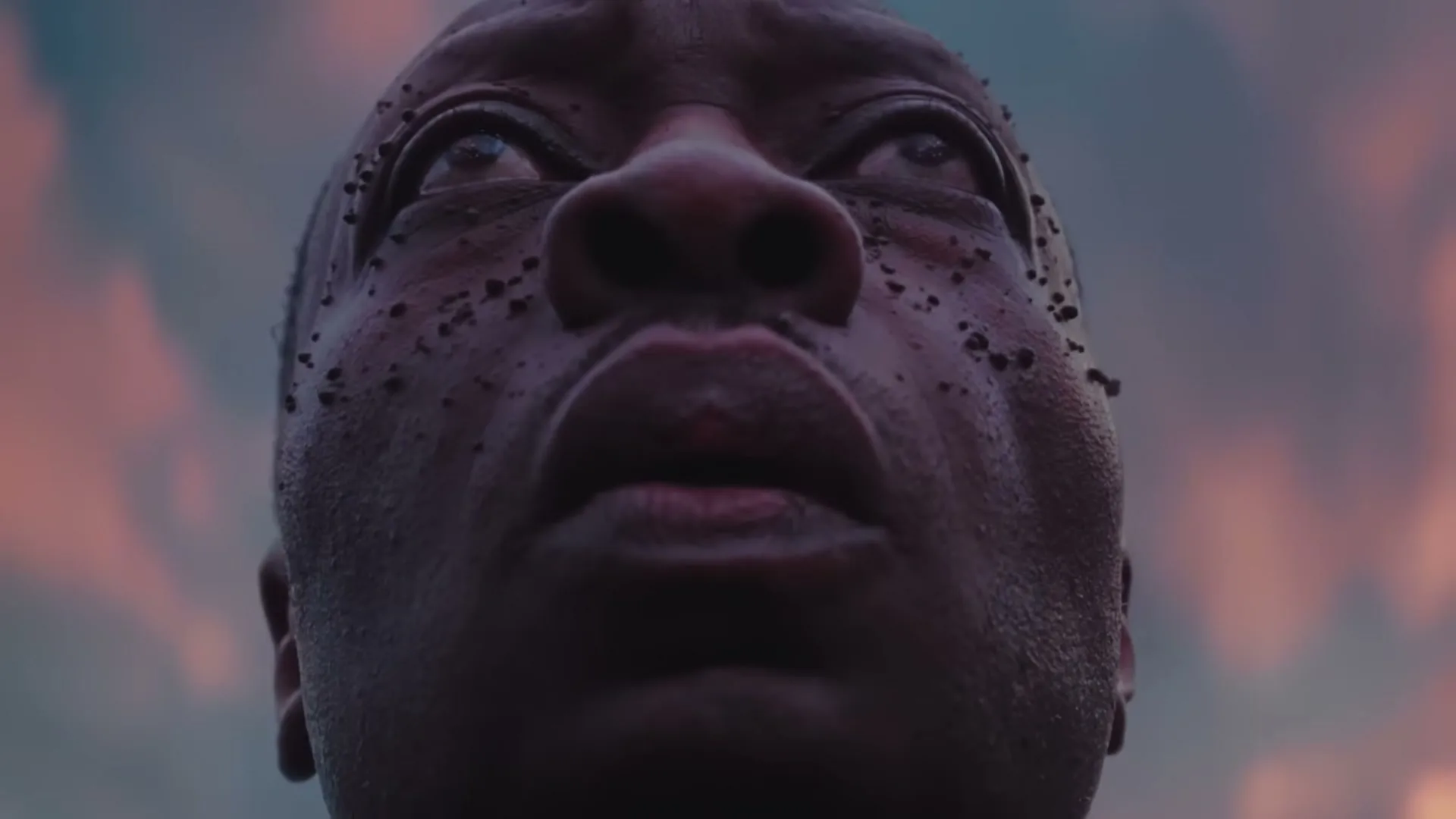 A man with dark skin and small dark particles on his face looks up with a serious expression against a blurred sky.
