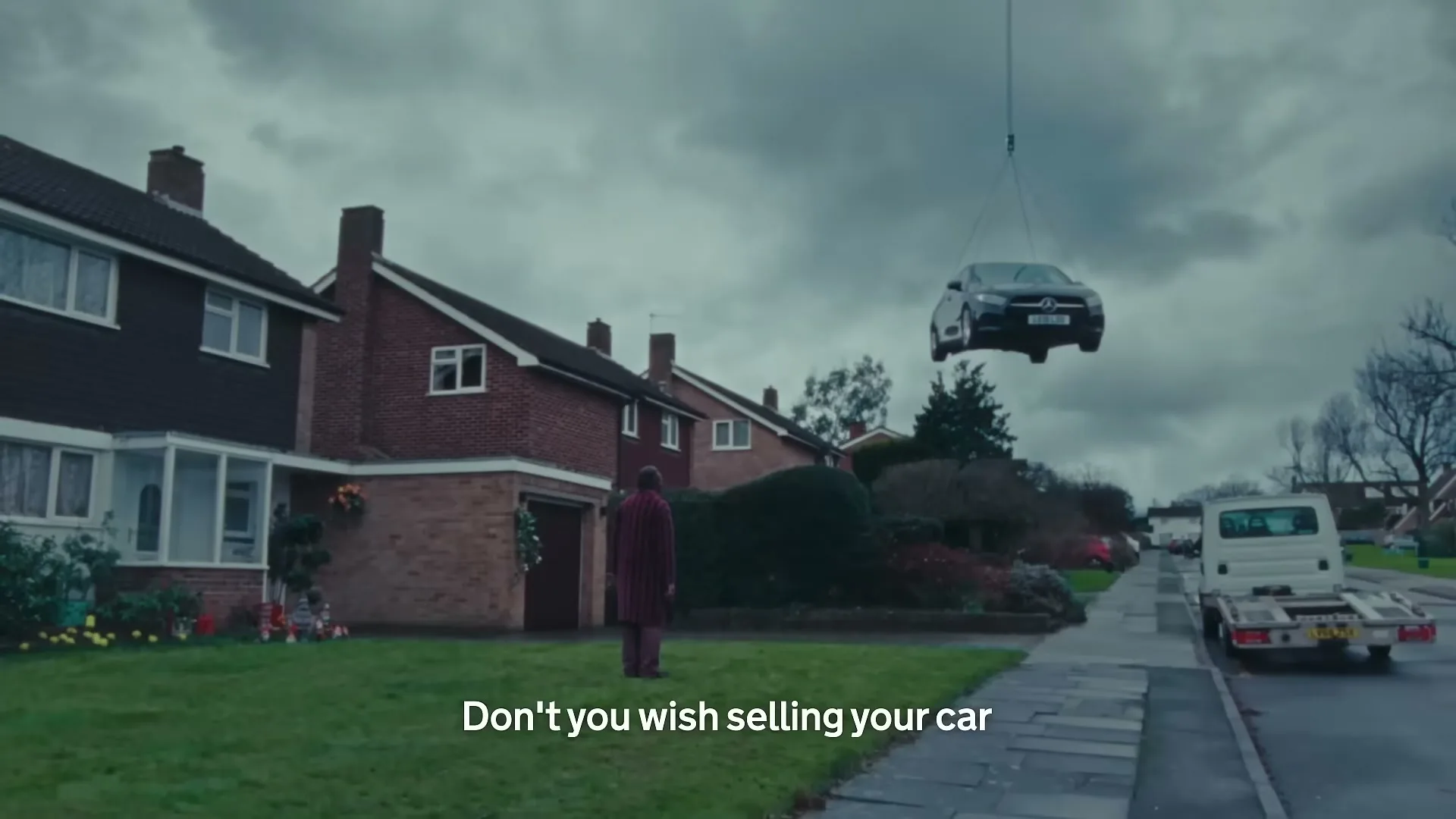 A man in a striped robe stands on a suburban lawn as a black car is lifted by a crane over a white flatbed truck on a cloudy day.