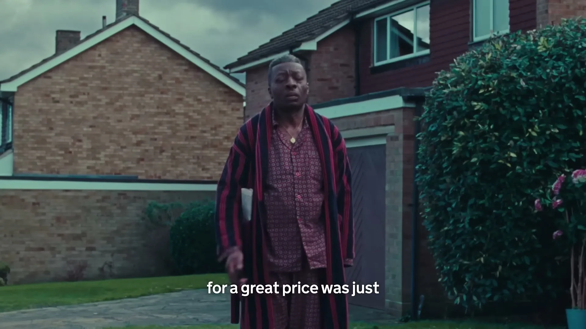 A man in his 50s, wearing pajamas and a robe, walks slowly in front of a suburban house, looking around with a confused expression.