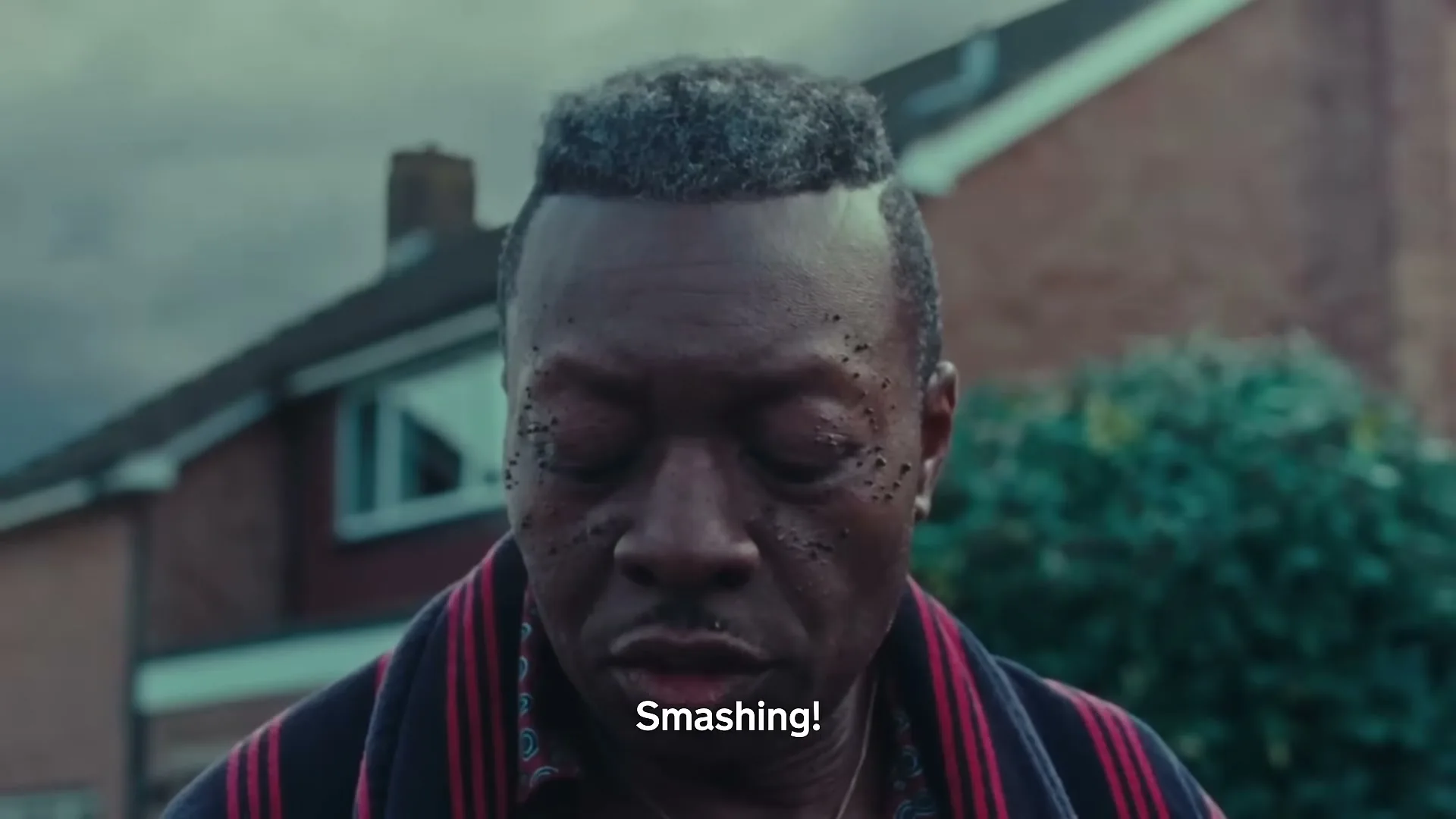A man with a unique hairstyle and facial markings closes his eyes, then opens them, looking down in a suburban setting.