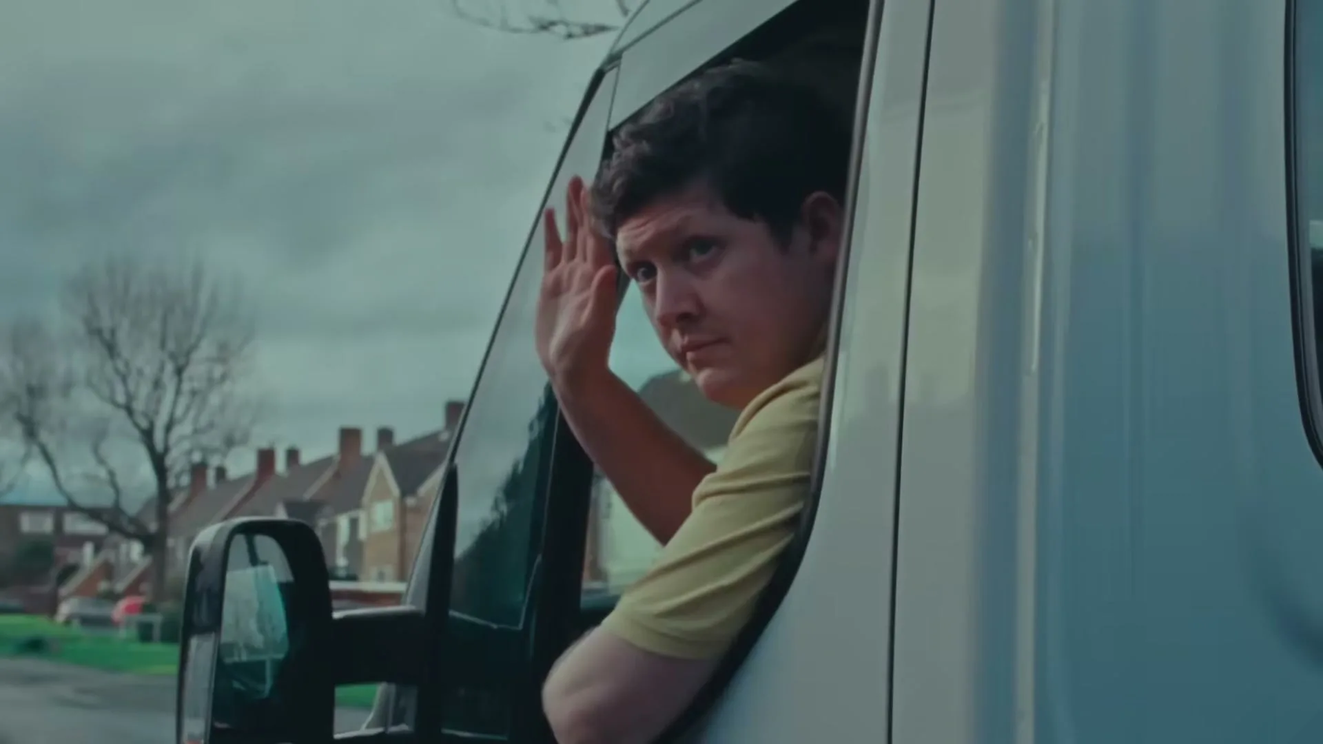 A man in a yellow shirt looks out the window of a white van and waves, on a cloudy day in a suburban neighborhood.