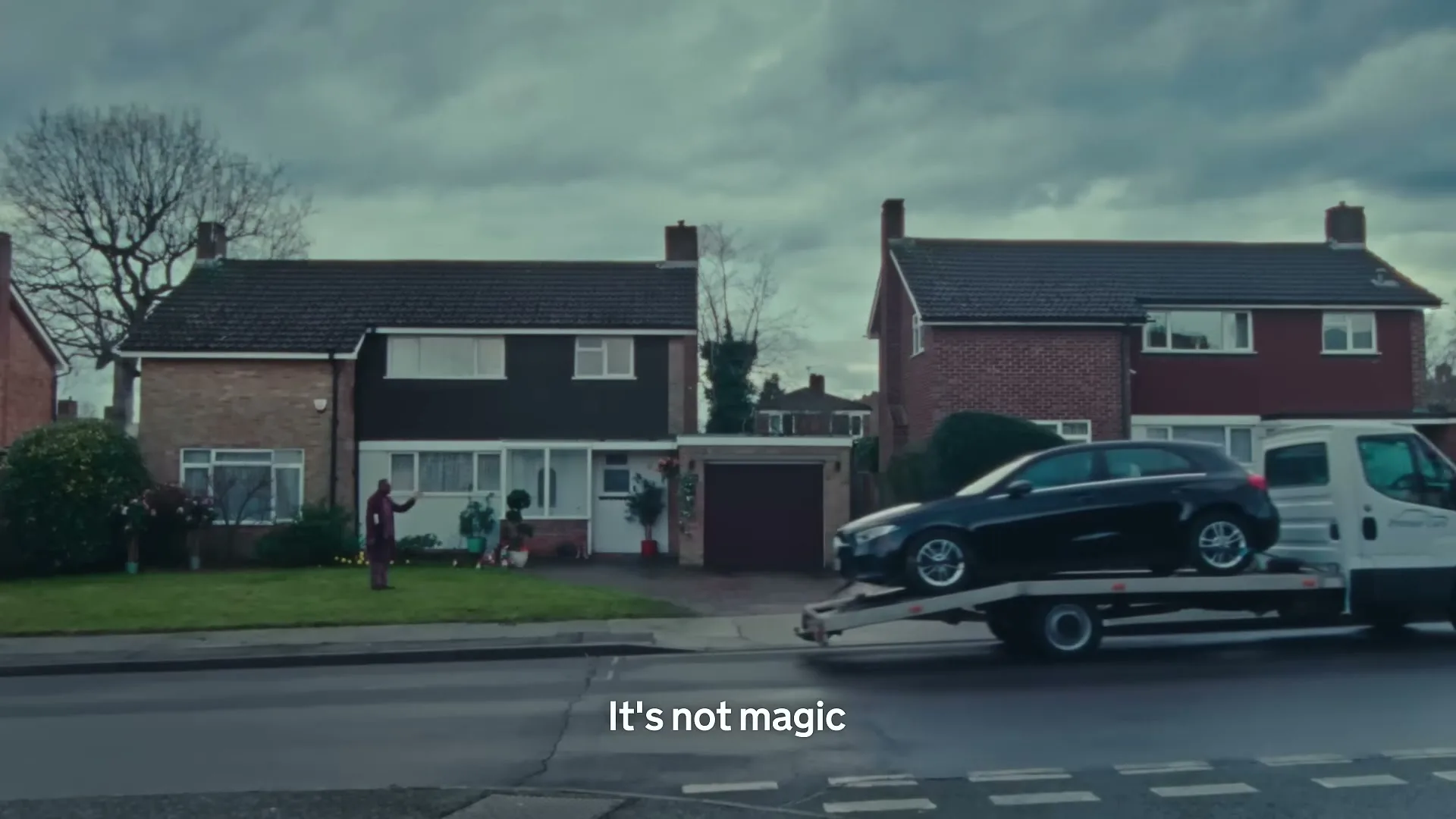 A man in a red suit stands on a lawn as a black car on a tow truck drives past, then a yellow smoke cloud appears.