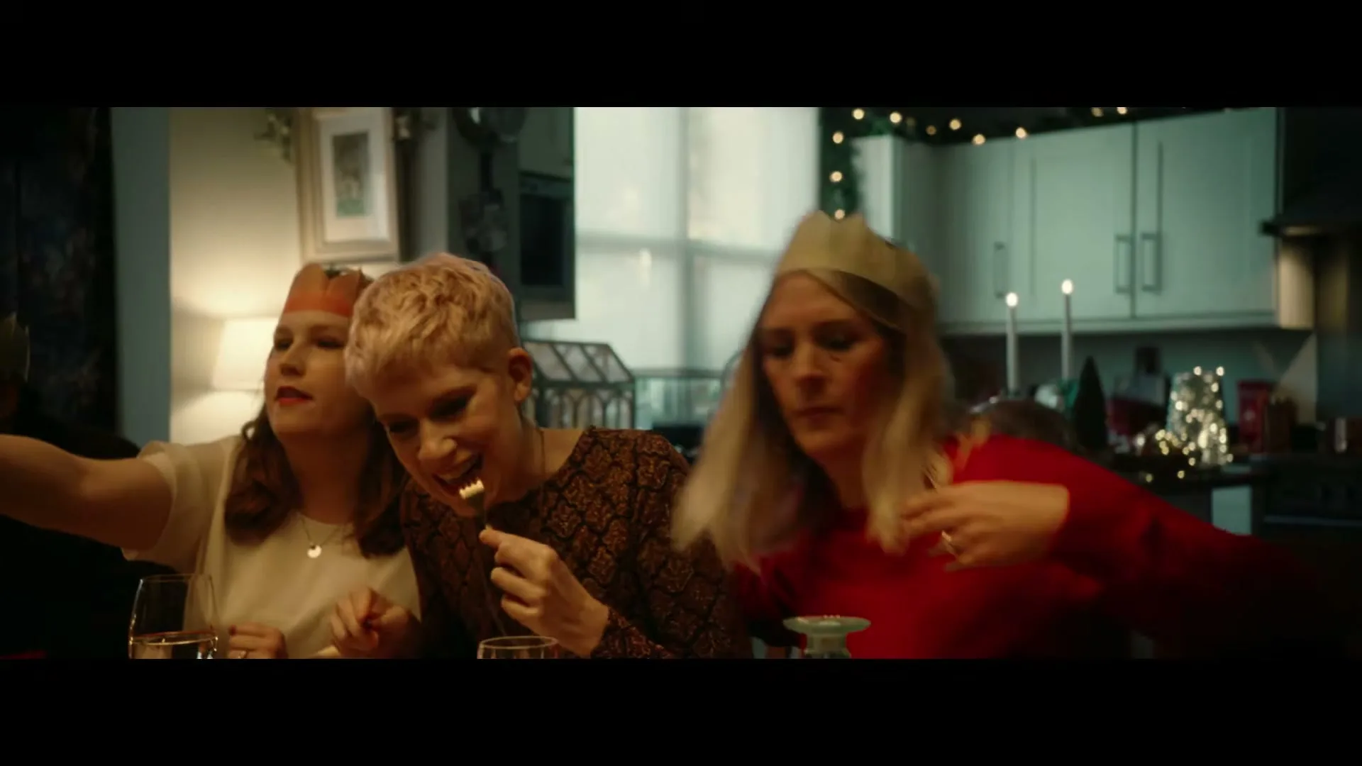 Three women, two wearing paper crowns, sit at a dining table, eating and talking during a Christmas dinner in a cozy home.
