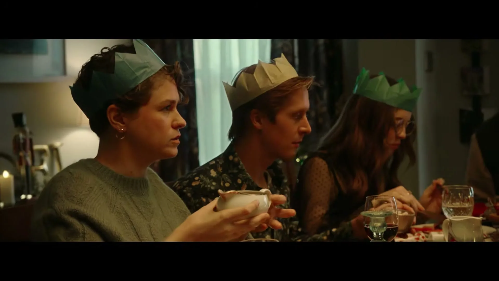 Three young adults wearing paper crowns sit at a dining table, eating and talking during a Christmas dinner.