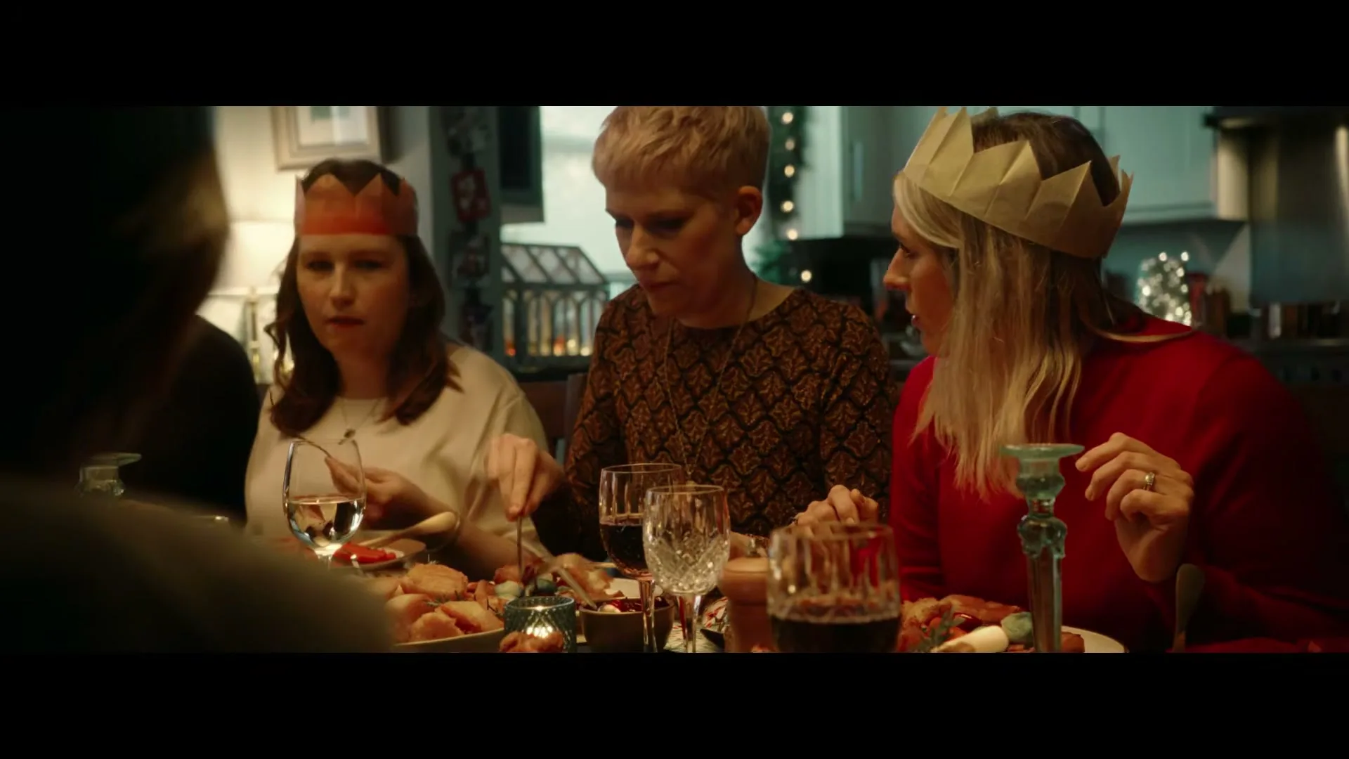 Three women wearing paper crowns are seated at a dining table, eating a Christmas meal in a cozy home.