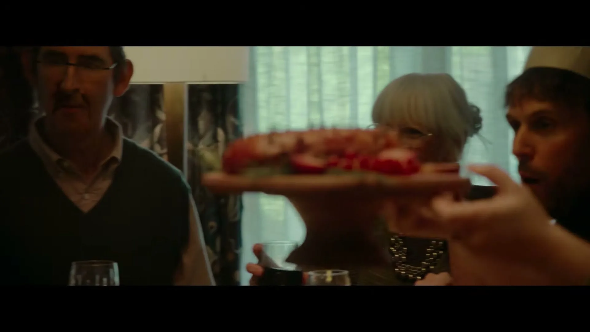 A family sits around a dining table, as a platter of red food is passed between hands, with a man and woman looking on.