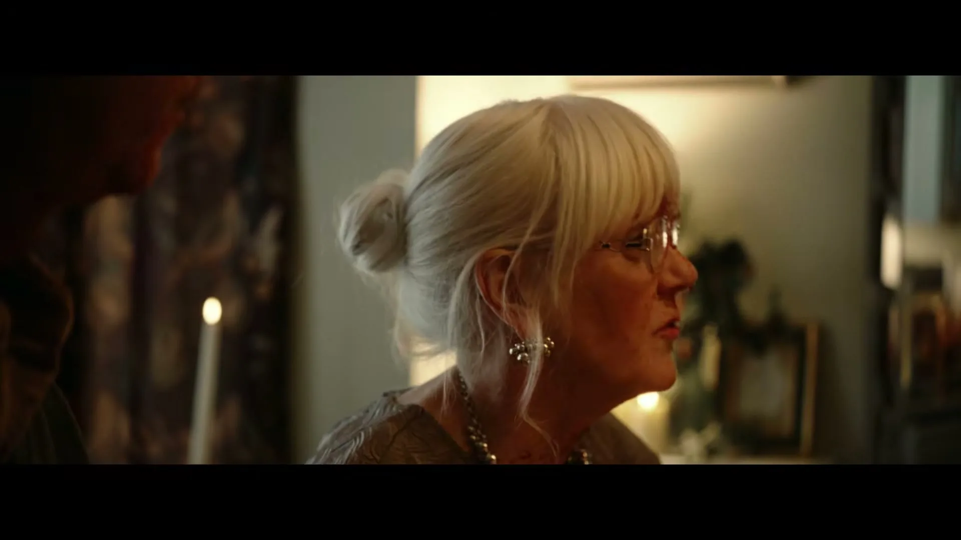An older woman with white hair and glasses, wearing a necklace, looks down at something off-screen while a man stands beside her in a dimly lit room.