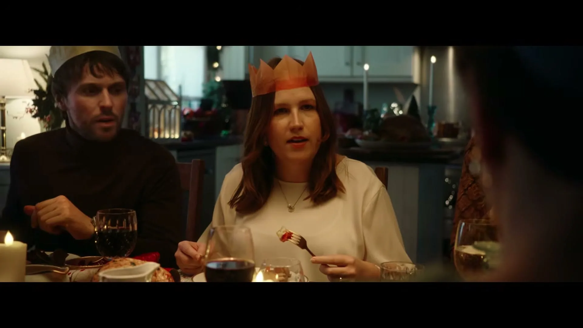 A woman in her 30s, wearing a white top and a paper crown, sits at a Christmas dinner table, holding a fork with food, looking at someone off-screen.