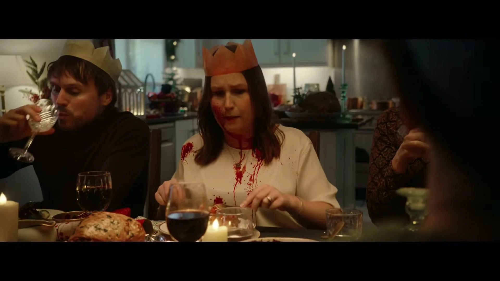 A woman in a paper crown, covered in blood, cuts food at a Christmas dinner table, while a man next to her drinks wine.