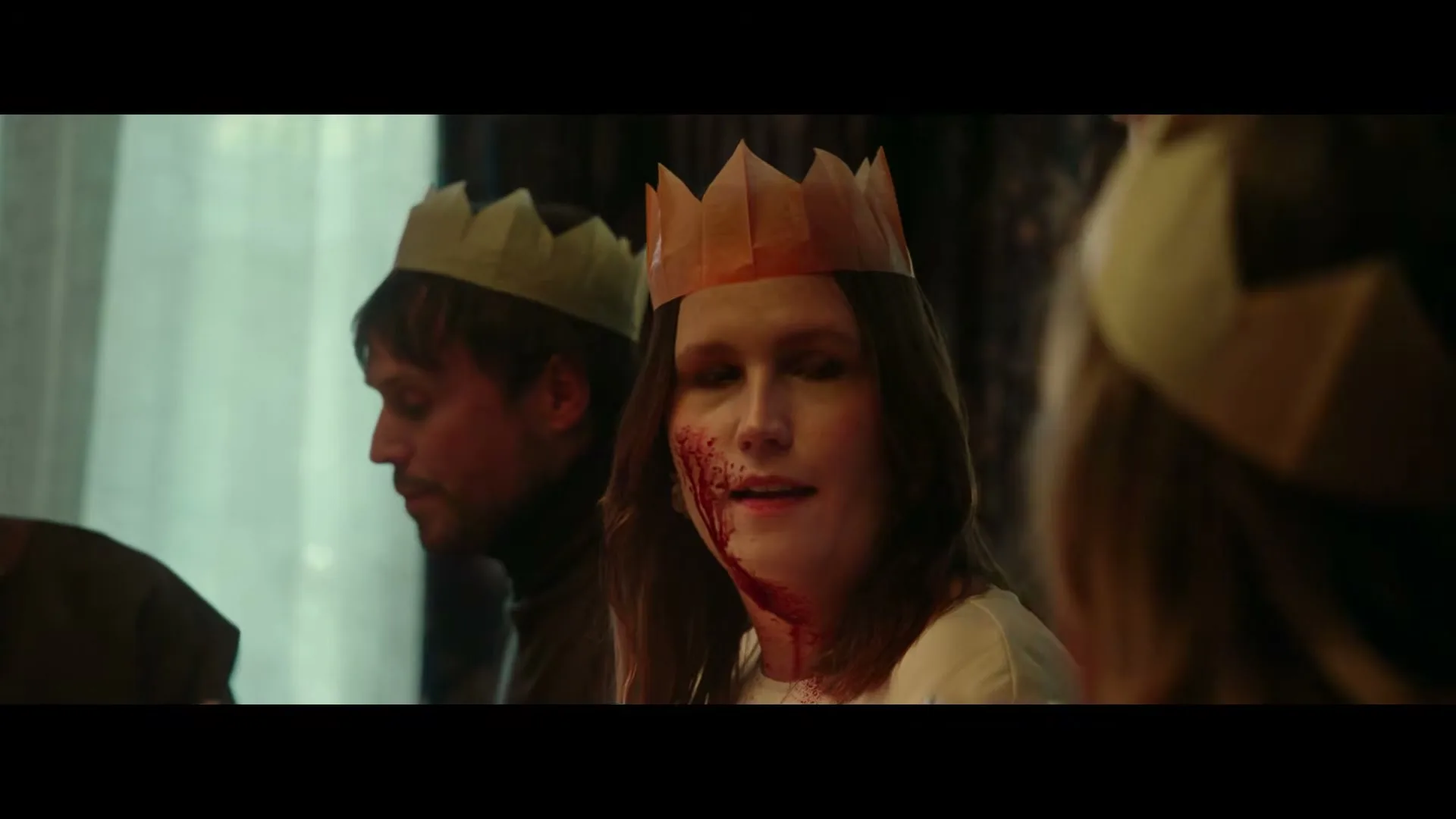 A woman with blood on her face and a paper crown looks at the camera while a man eats in the background at a Christmas dinner.