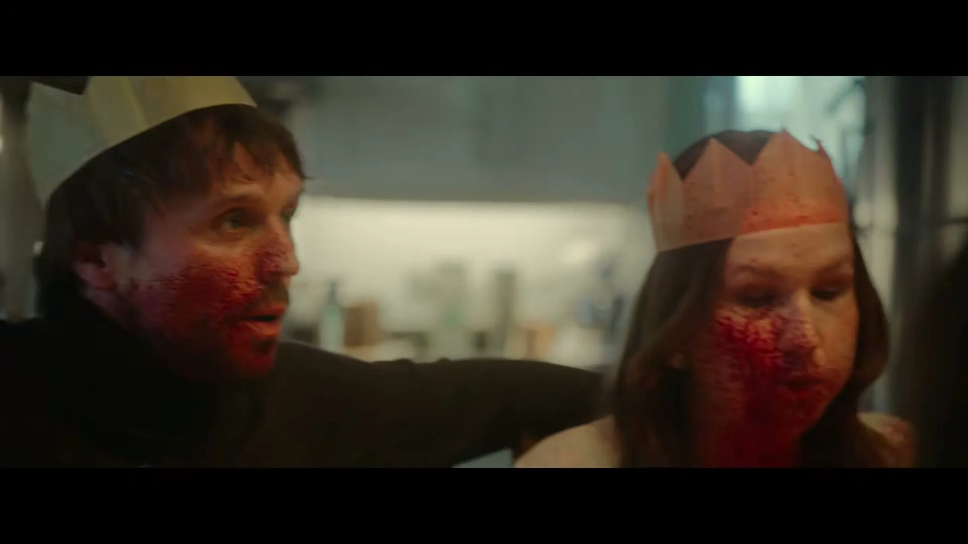 A man and a woman, both with blood splattered on their faces and wearing paper crowns, look shocked and scared in a dimly lit kitchen.