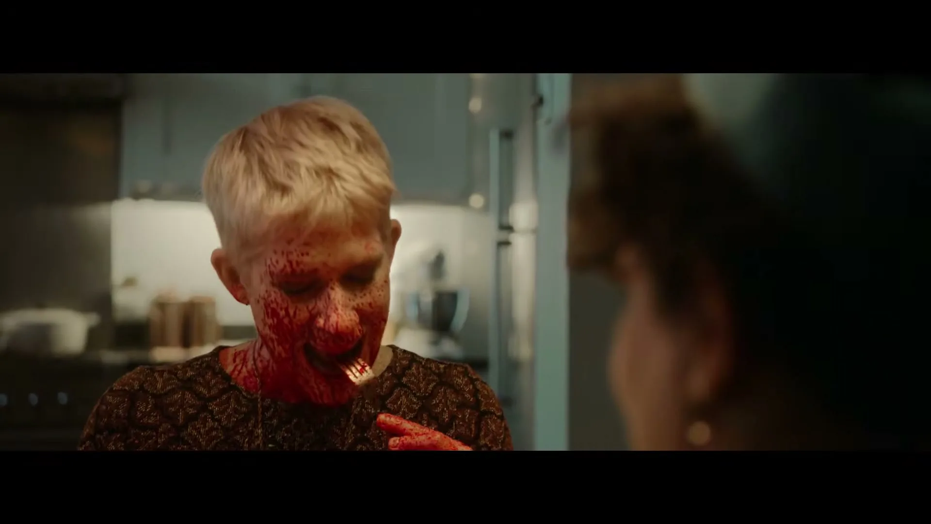A woman with short blonde hair and a blood-splattered face looks down, then up, then down again, in a modern kitchen.