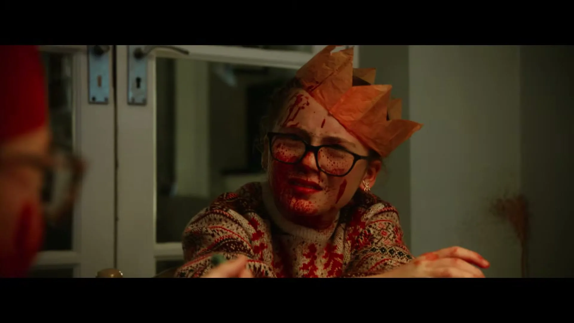 A young woman, covered in blood and wearing a paper crown, looks distressed while sitting at a table indoors.