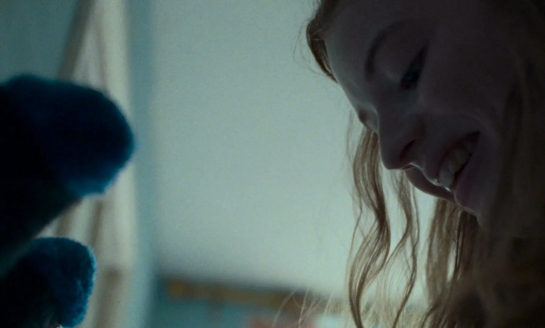 A young girl with long red hair looks down and smiles, playing with a blue puppet in a dimly lit room.
