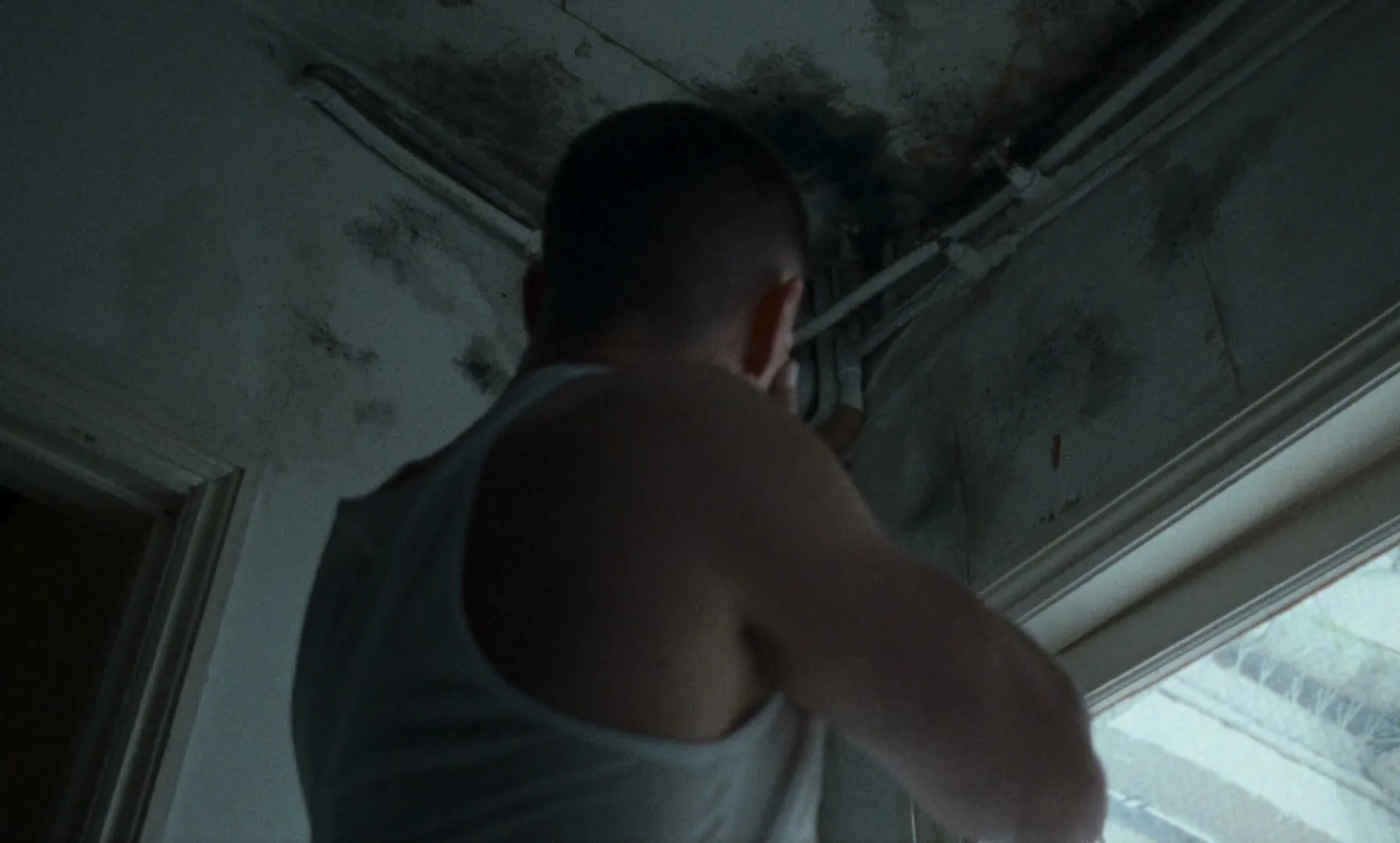 A man in a white tank top sprays a black bottle at a moldy ceiling in a dilapidated room, looking concerned.