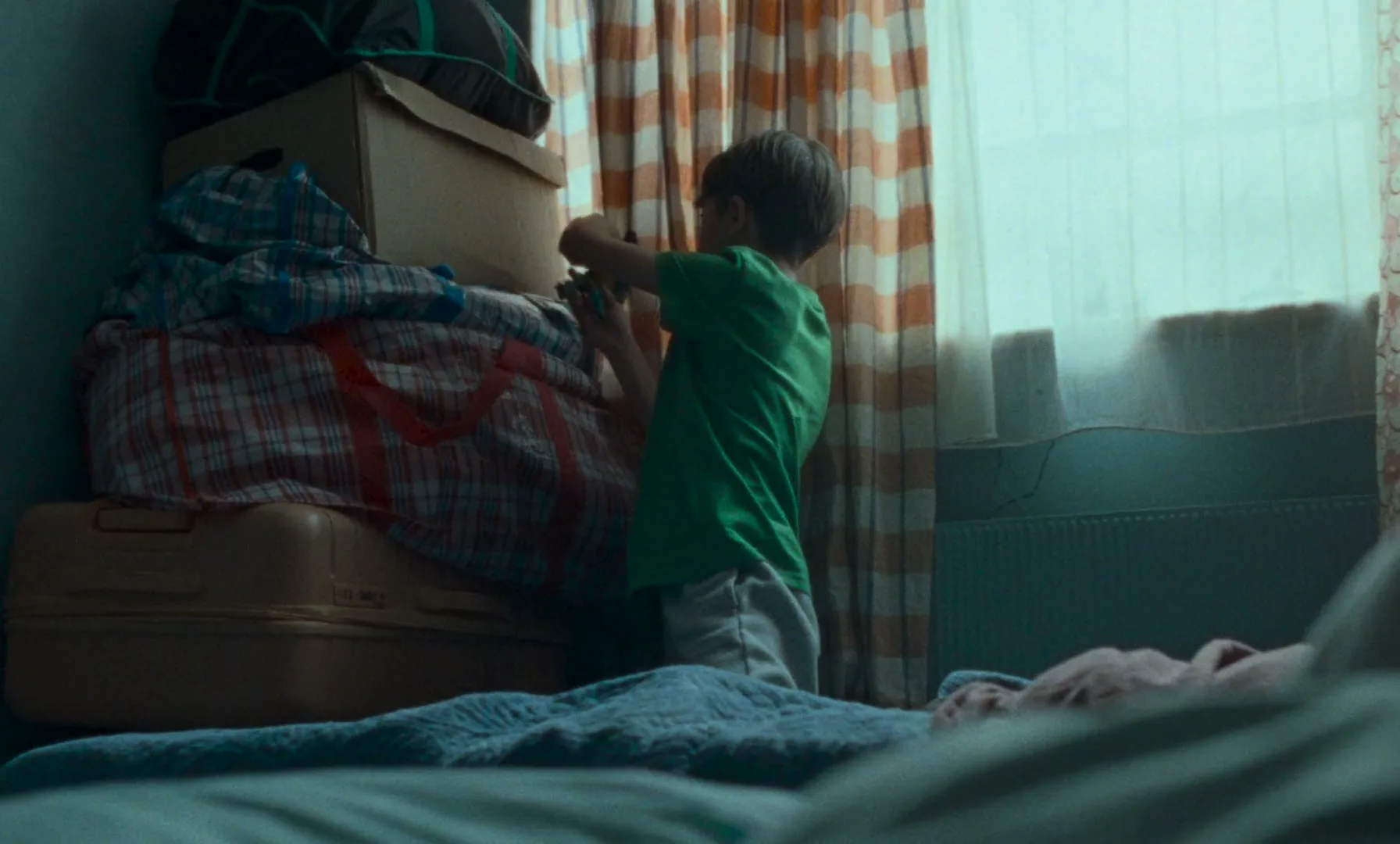 A young boy in a green shirt plays with a toy next to a stack of boxes and luggage in a dimly lit room.