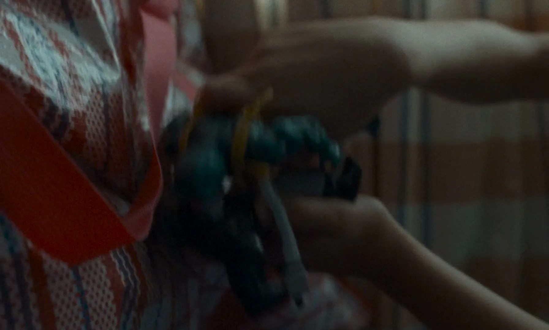 A child's hands hold a green action figure, partially obscured by a patterned bag, in a dimly lit interior.