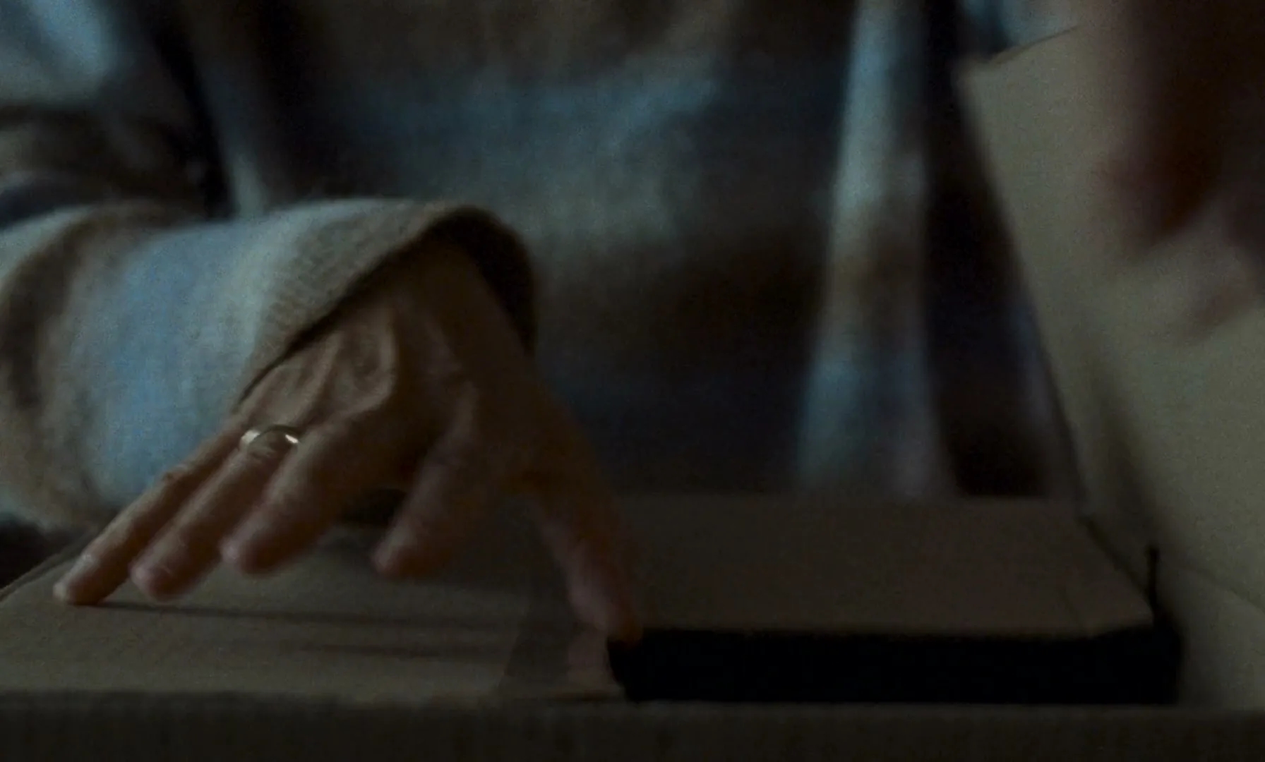 A person's hands are seen opening a cardboard box with a knife, revealing a dark object inside, in a dimly lit interior.
