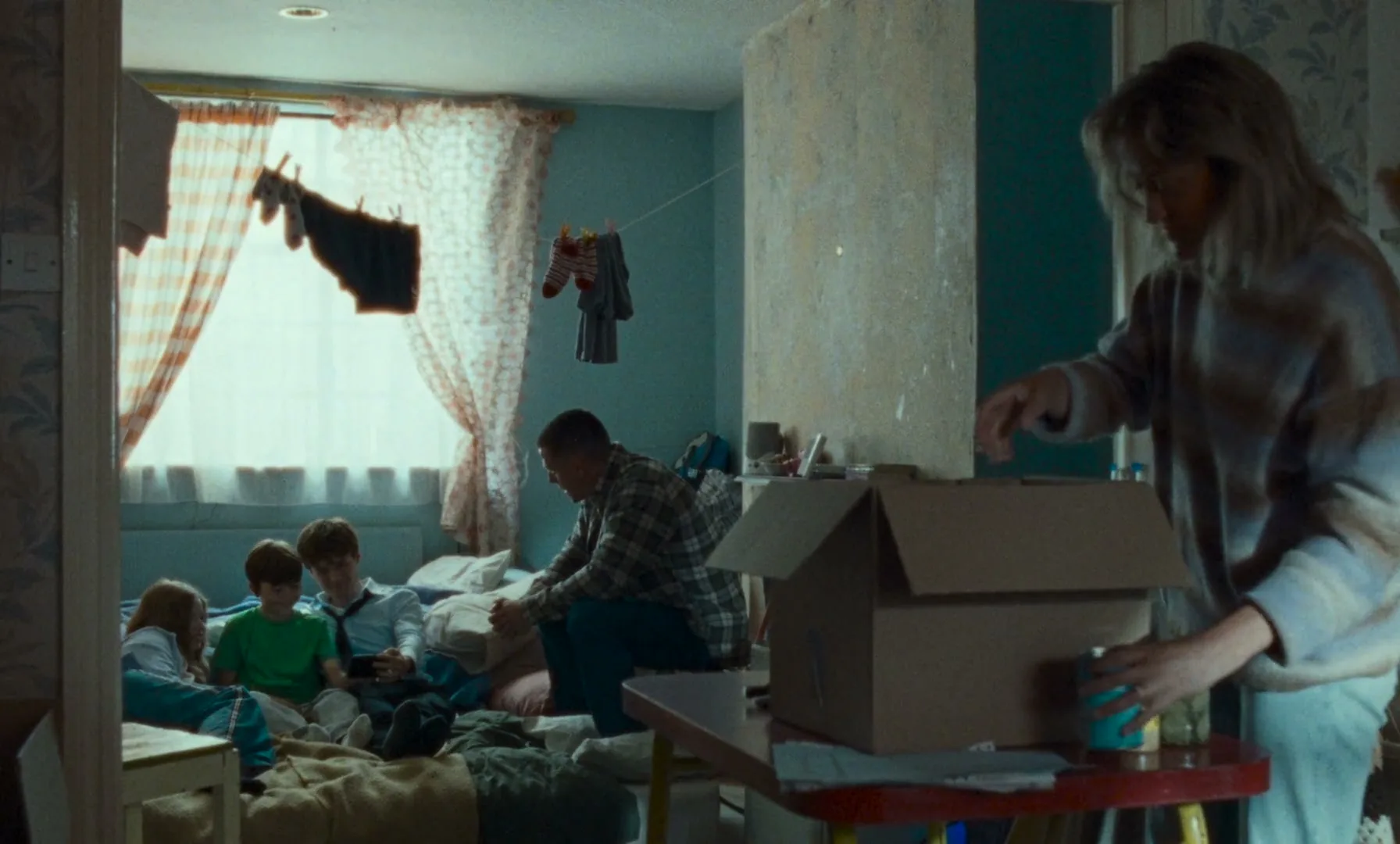 A family of five is in a cramped, messy room, with a woman packing items into a box while a man and three children sit on a bed.